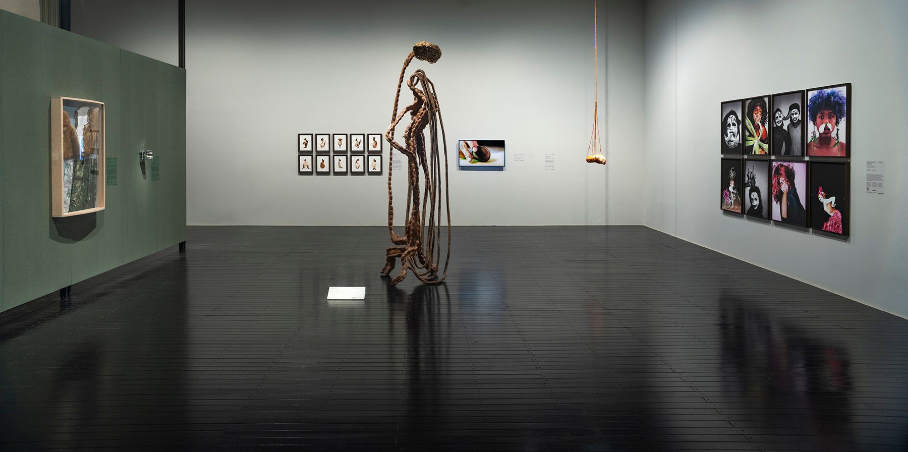 A modern art gallery with dark, reflective floors, light walls, a tall twisted sculpture, multiple framed artworks including portraits, a digital display, and a wooden swing hanging from the ceiling.