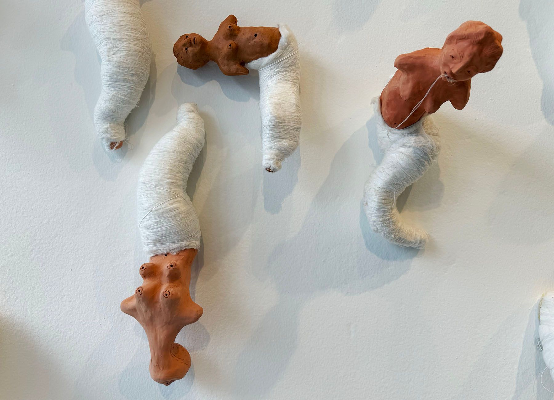 Abstract sculptures of reddish-brown clay forms partially wrapped in white fabric, displayed on a white wall.