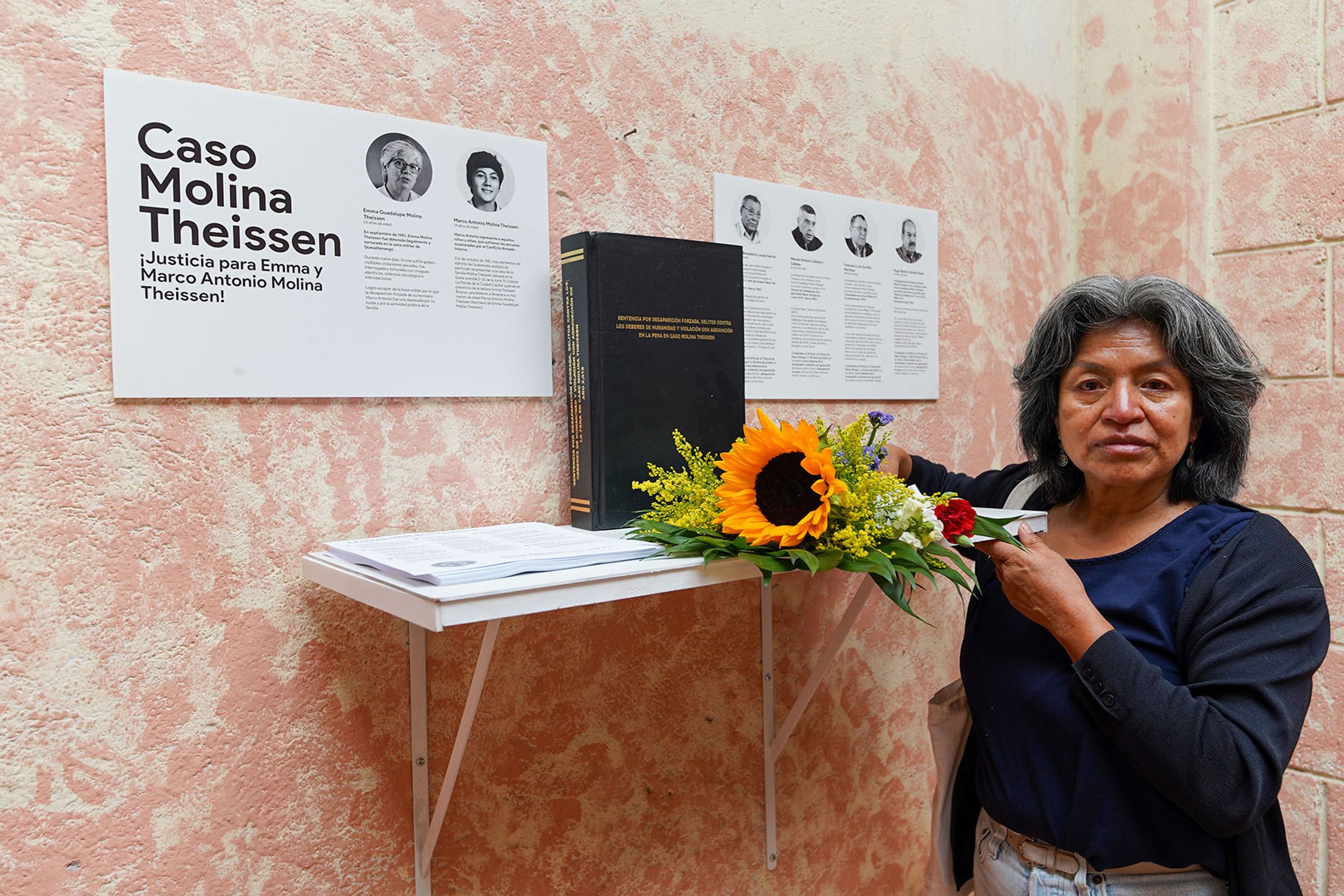 A woman stands by a display about the "Caso Molina Theissen" with a book, a sunflower arrangement, and posters, holding a red flower.