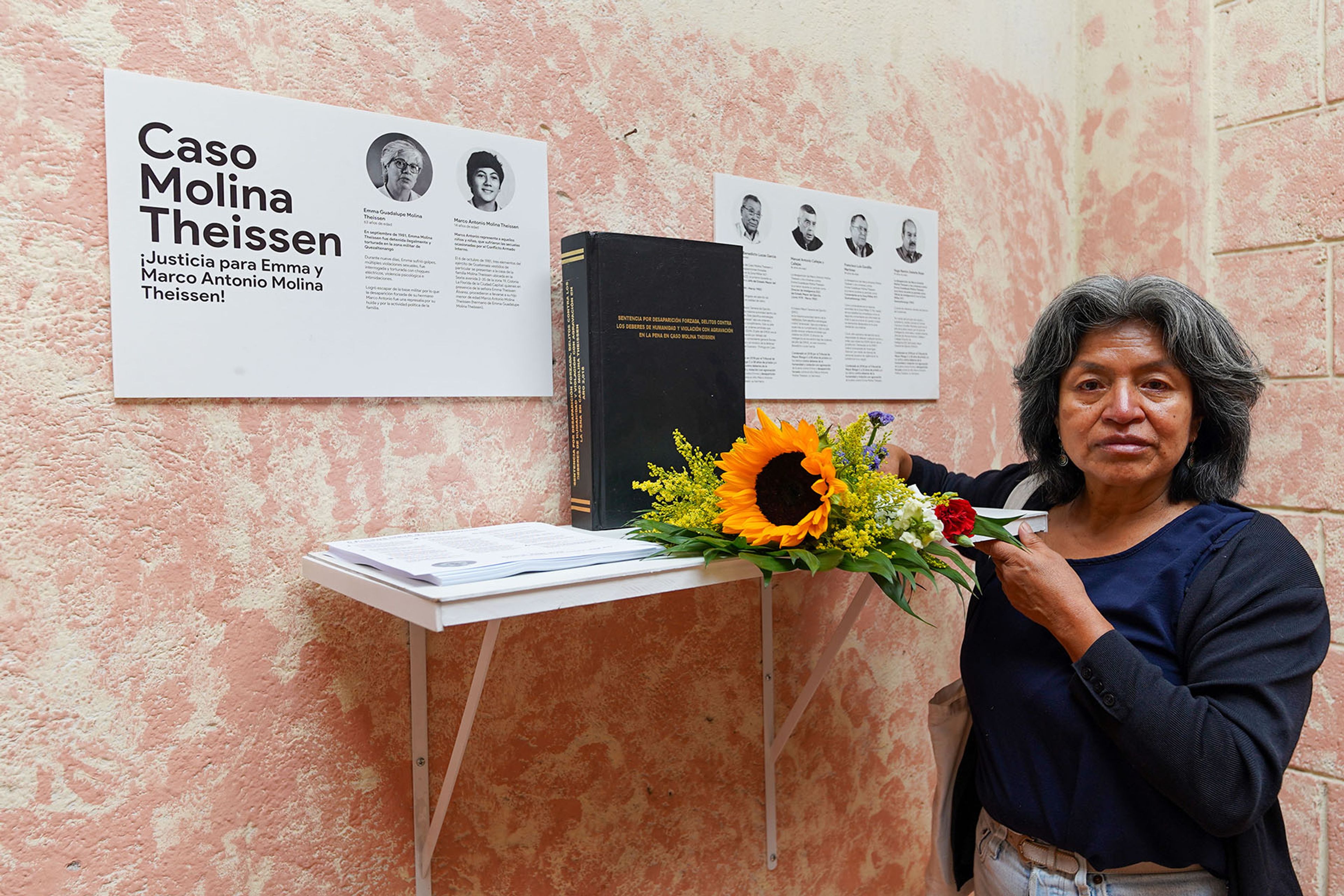 A woman stands by a display about the "Caso Molina Theissen" with a book, a sunflower arrangement, and posters, holding a red flower.