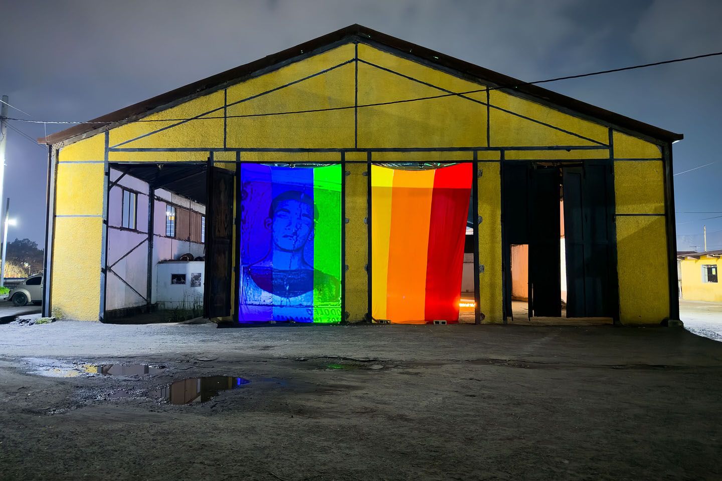 A yellow warehouse at night with two doorways displaying a Pride flag, featuring a person's face in blues and purples on the left and vertical green, yellow, orange, and red stripes on the right.