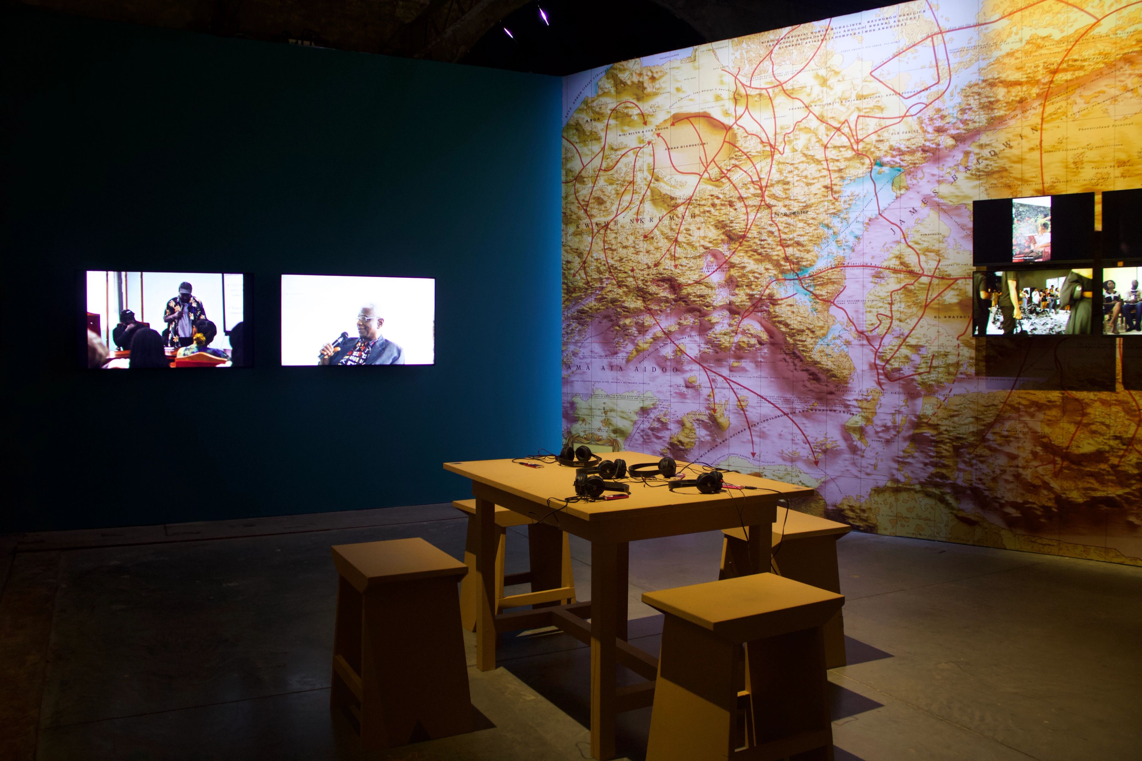 A multimedia exhibit featuring a dark teal wall with two video screens, an adjacent wall with a large projected topographical map and six smaller screens, and a wooden table with headphones and stools.