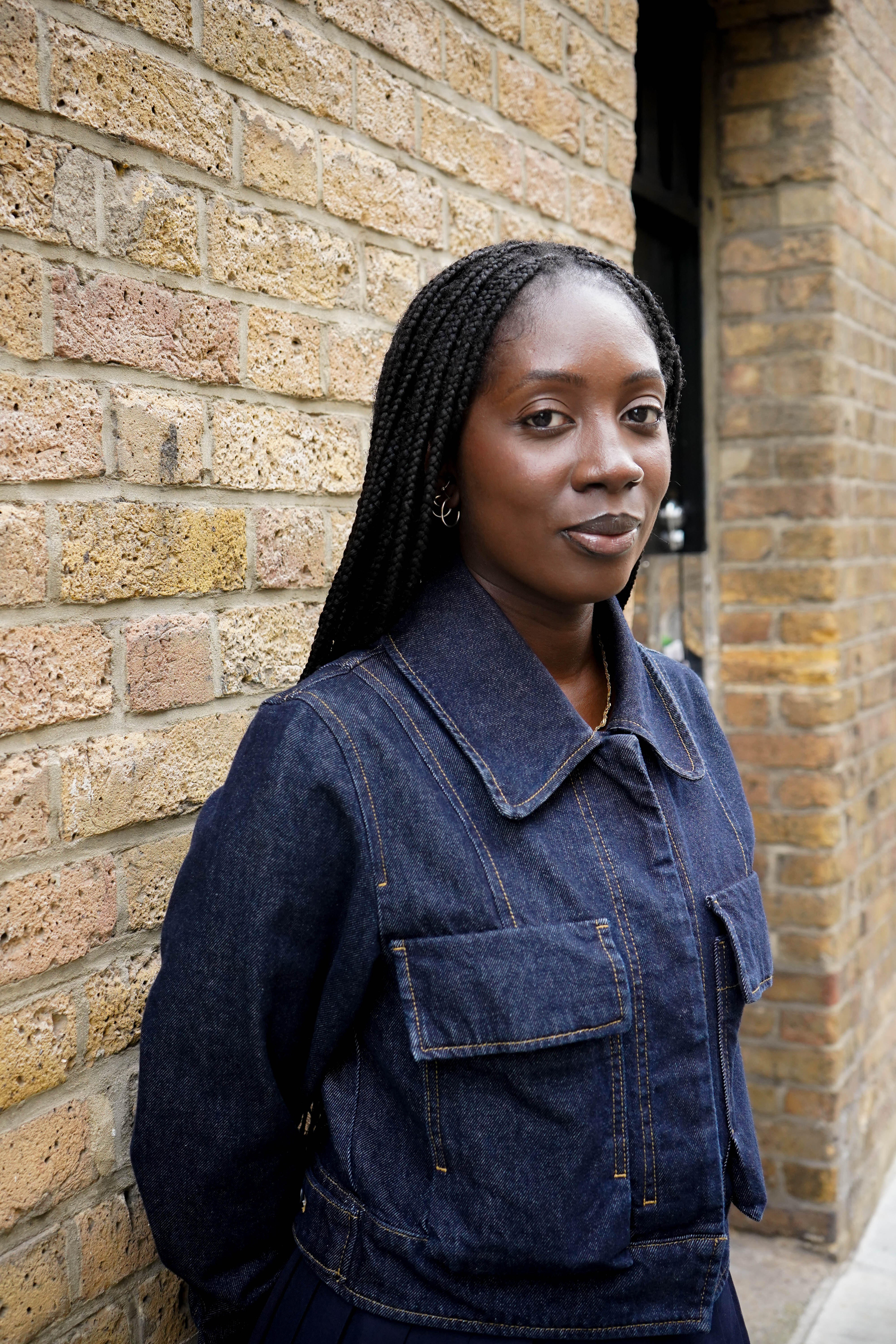 A young Black woman with braided hair wearing a denim jacket stands against a brick wall.