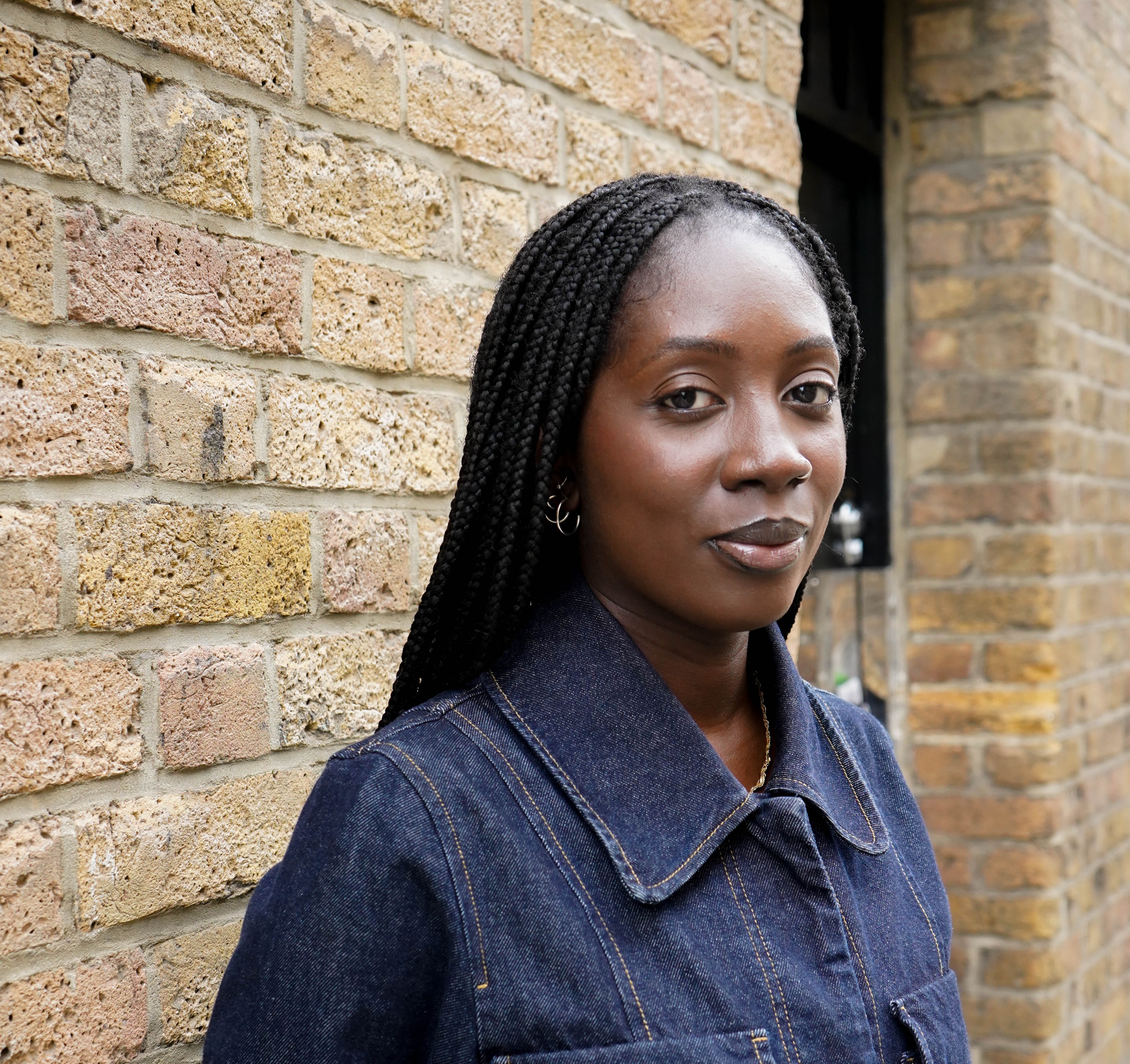 A young Black woman with braided hair wearing a denim jacket stands against a brick wall.