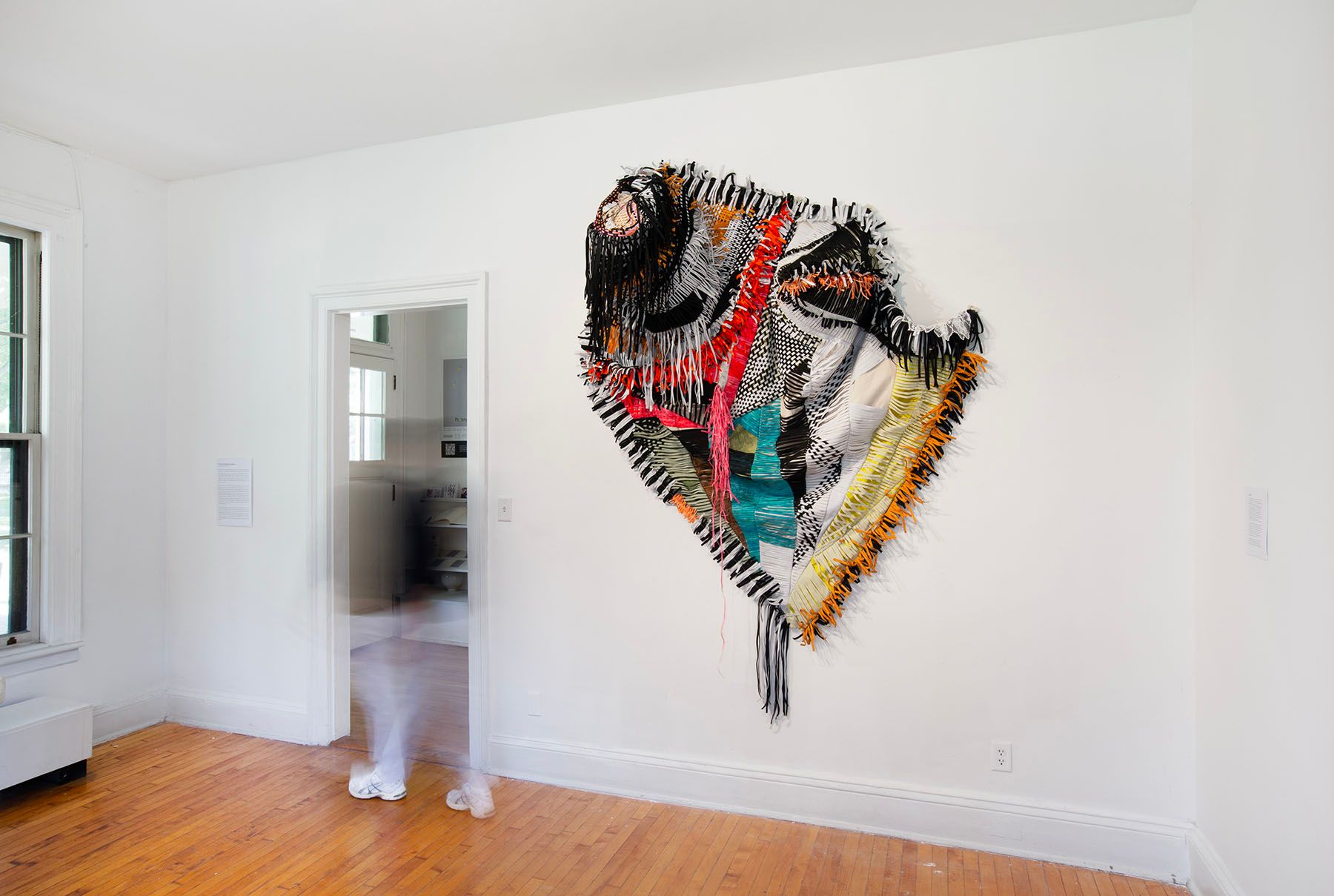 A bright white room features a large, multi-colored, shield-like textile artwork on the wall, with a blurred figure walking through an open doorway.