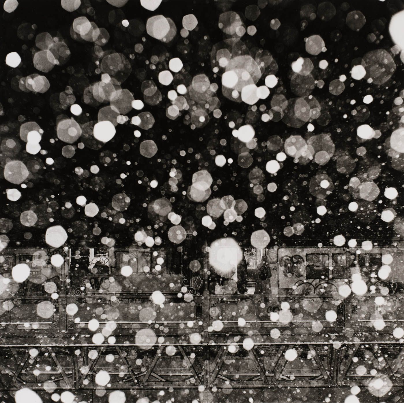 Black and white photo of illuminated falling snow over a blurry dark urban scene with a bridge.