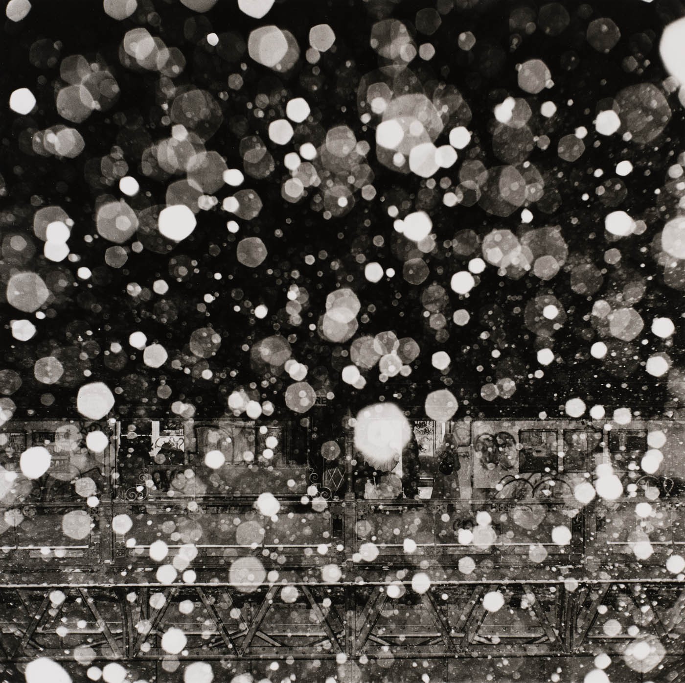 Black and white photo of illuminated falling snow over a blurry dark urban scene with a bridge.