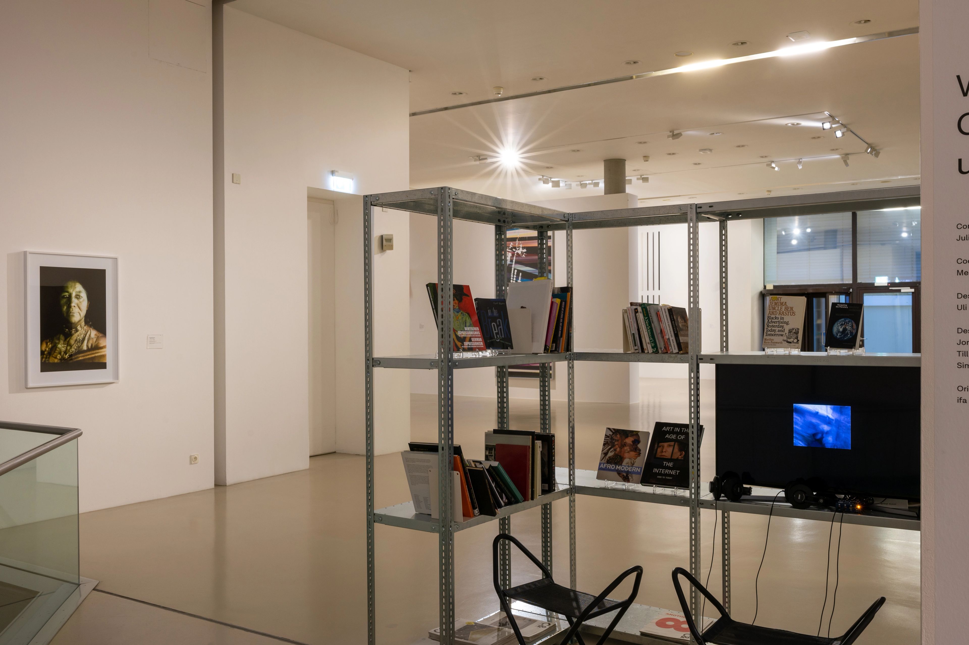 An L-shaped metal shelving unit with books and a TV playing a video, two chairs with headphones, and a framed figurative artwork on a white gallery wall.