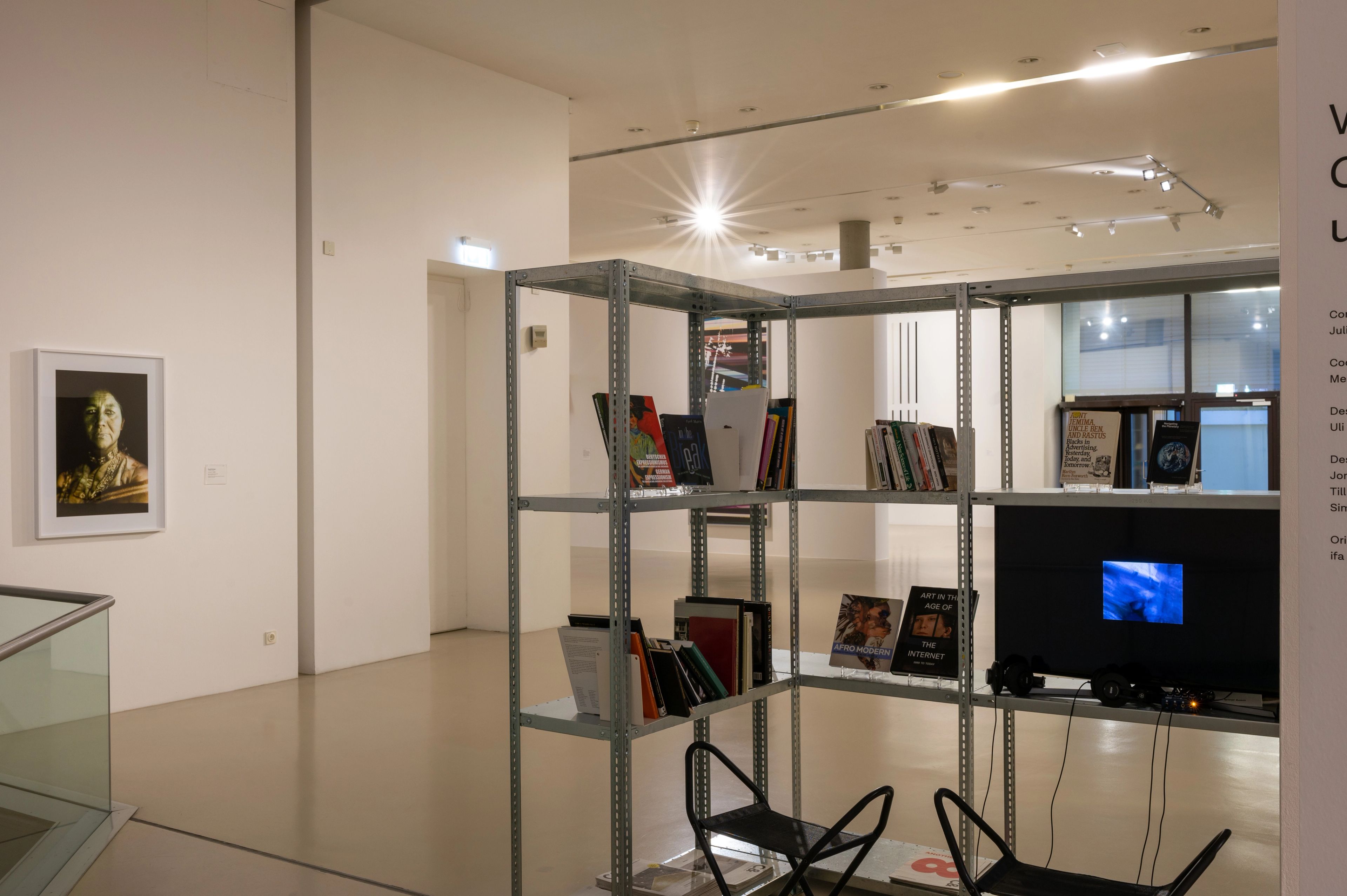An L-shaped metal shelving unit with books and a TV playing a video, two chairs with headphones, and a framed figurative artwork on a white gallery wall.