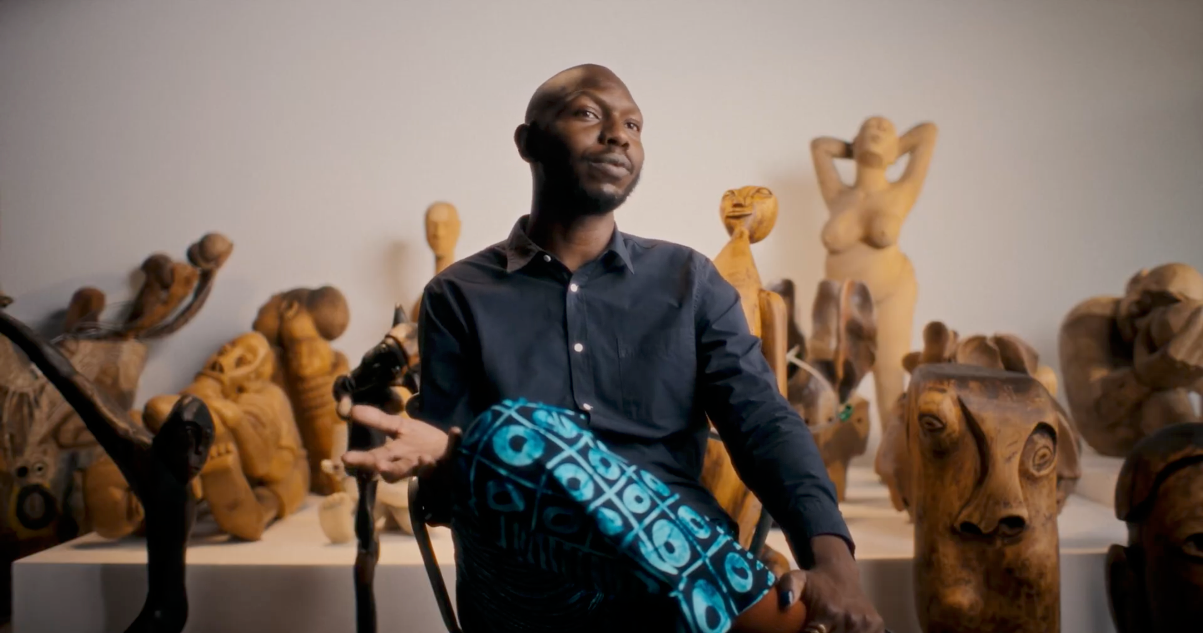 A Black man sits among numerous wooden sculptures in an art studio.