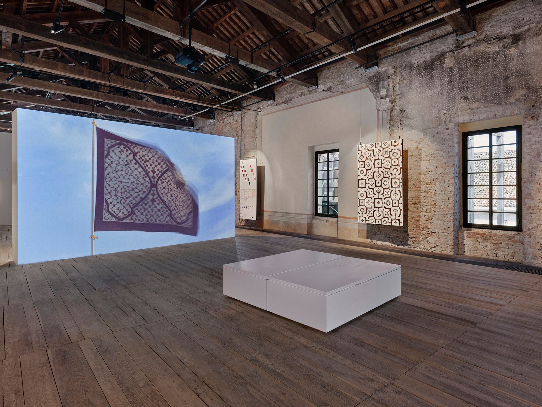 An art exhibition featuring a large projected flag, patterned wall art, and a white display platform in a rustic room.