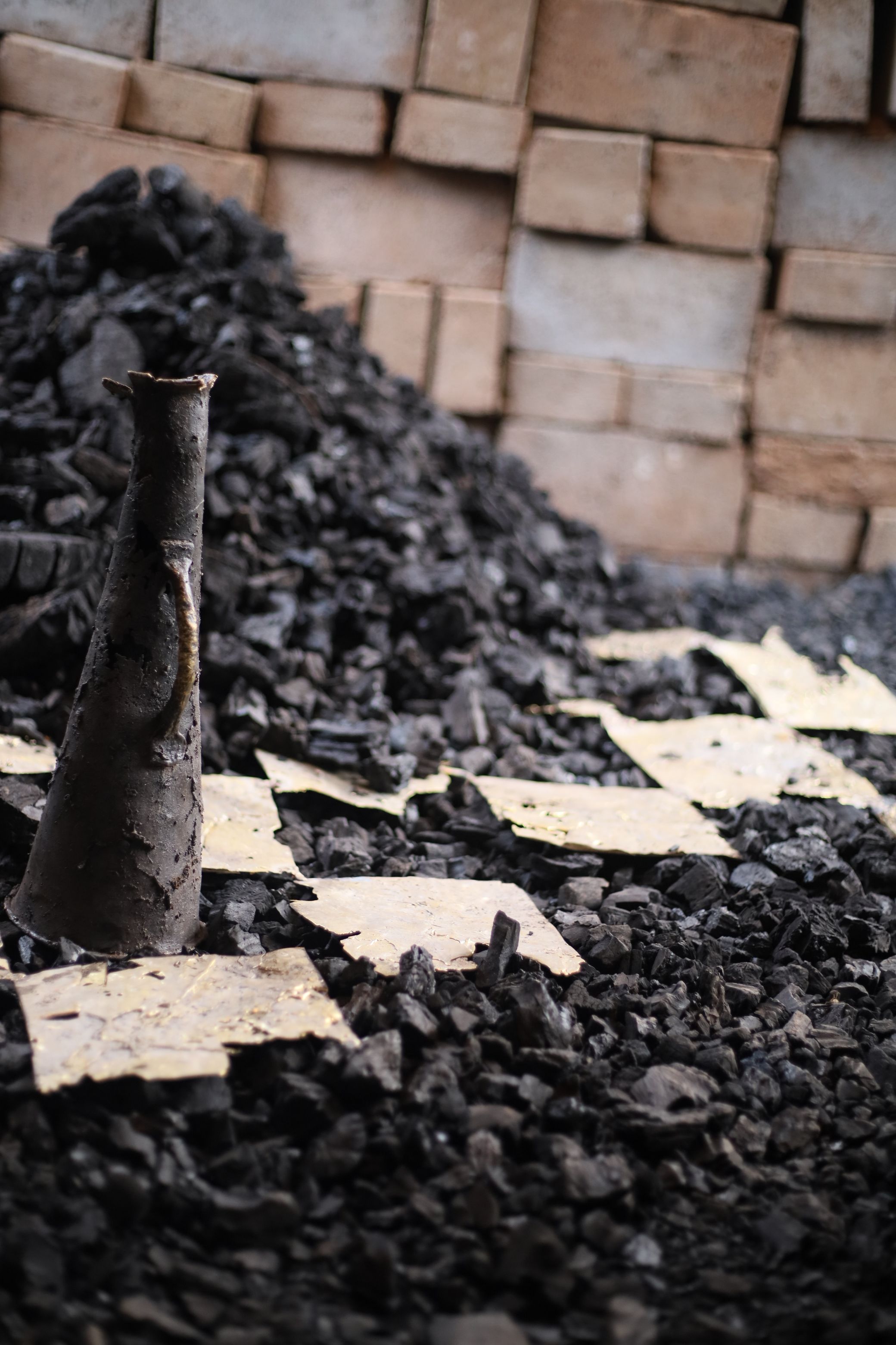 A dark conical object stands upright in a pile of charcoal and light fragments, with a brick wall in the background.
