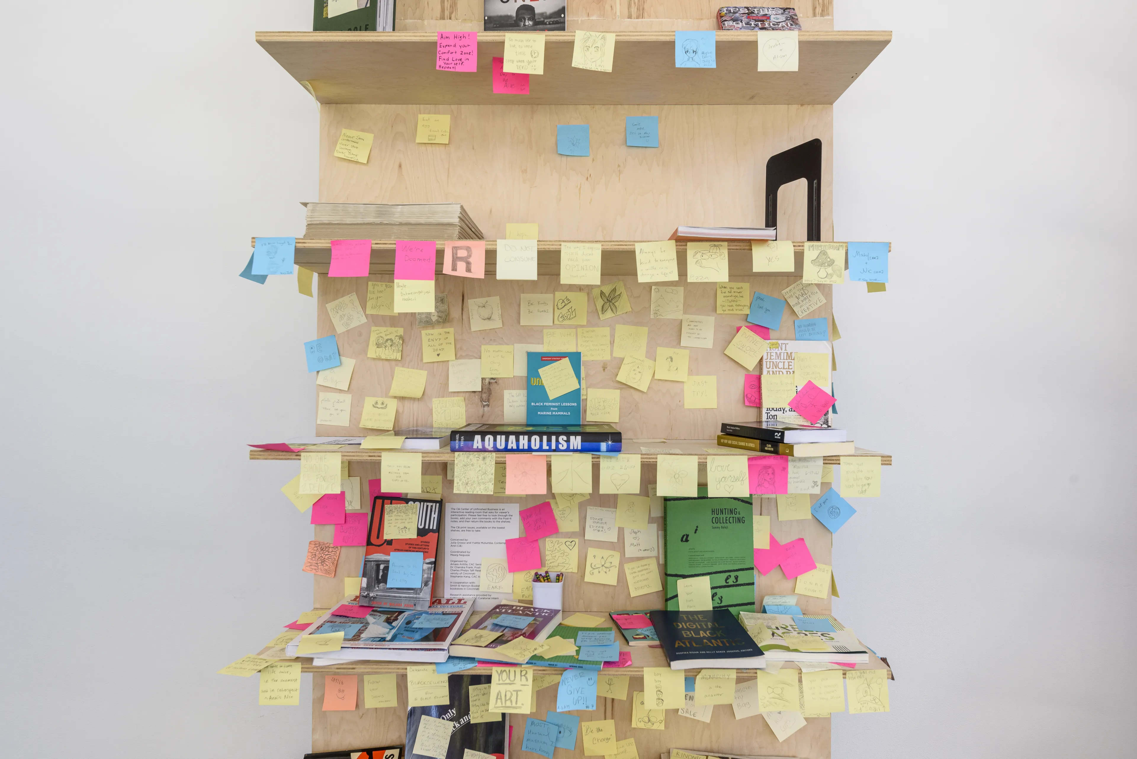 A wooden bookshelf heavily covered with yellow, pink, and blue sticky notes, displaying several books.