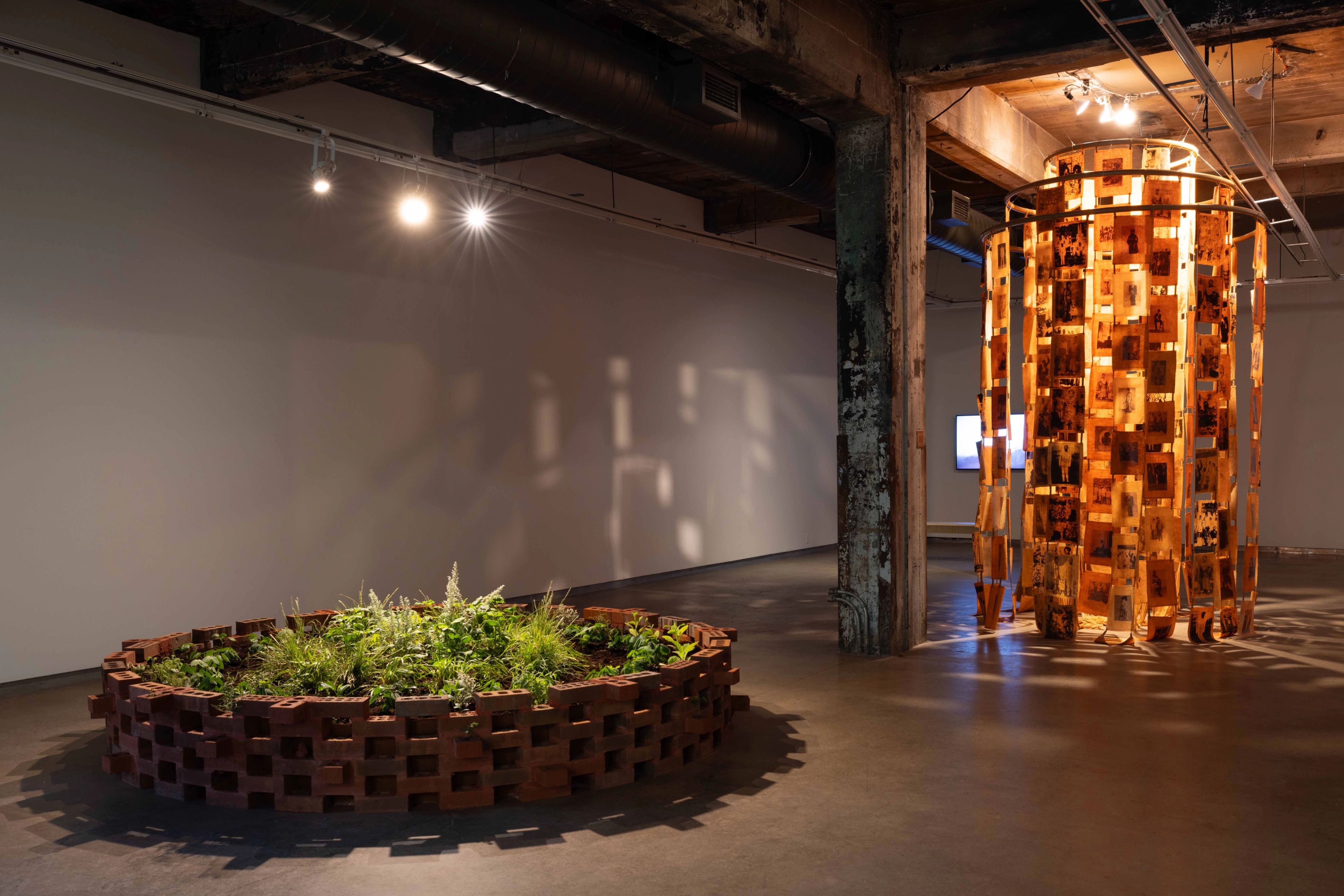 An art installation with a circular brick planter of green plants and a tall, glowing cylindrical sculpture of illuminated panels.