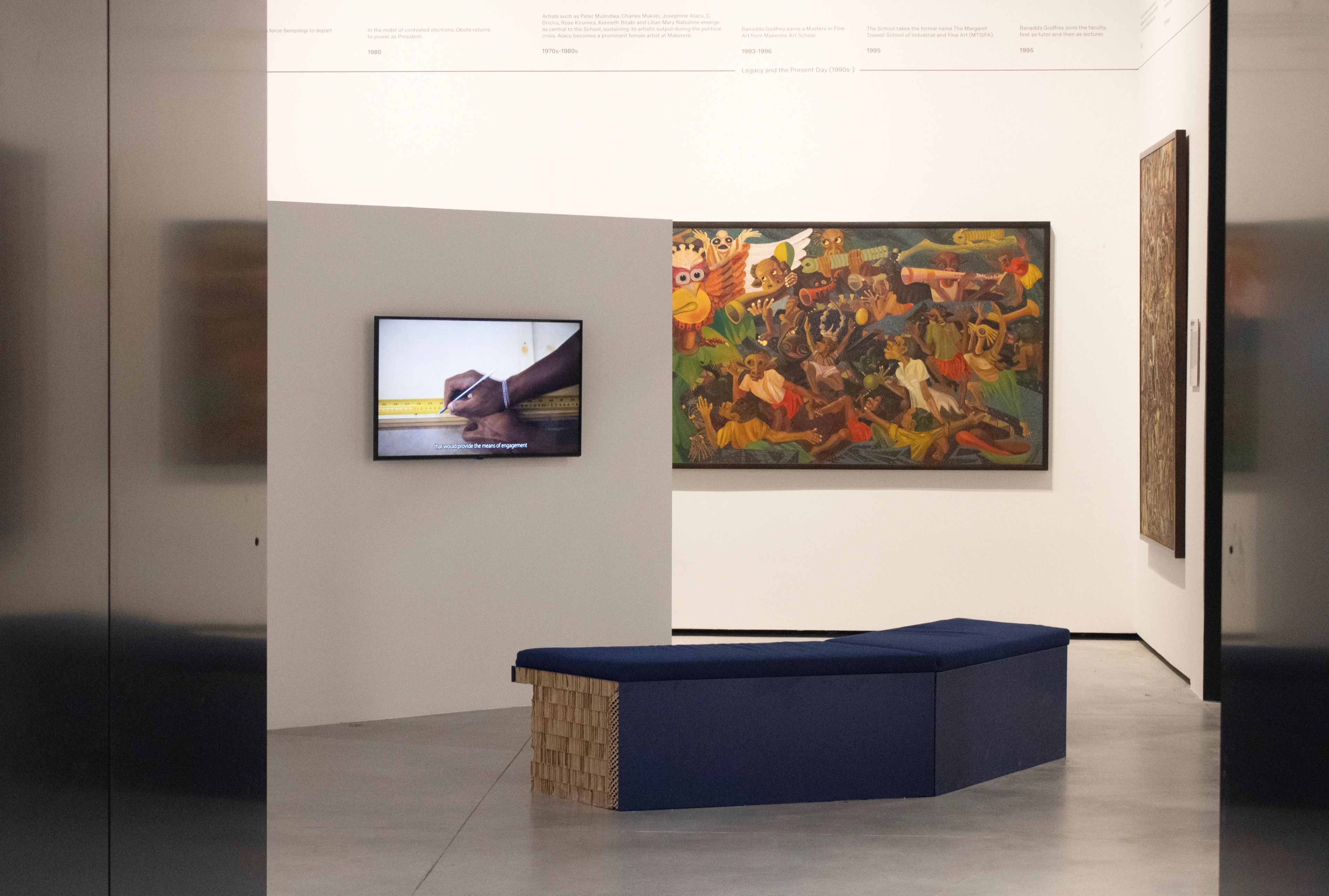 Art gallery with a large colorful painting, a screen showing hands measuring, and a long blue bench.