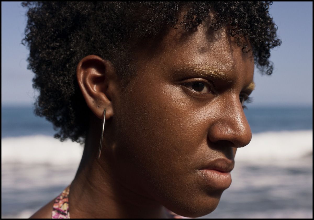 Close-up of a person with dark skin, curly hair, and bleached eyebrows looking right with a pensive expression, against an ocean backdrop.