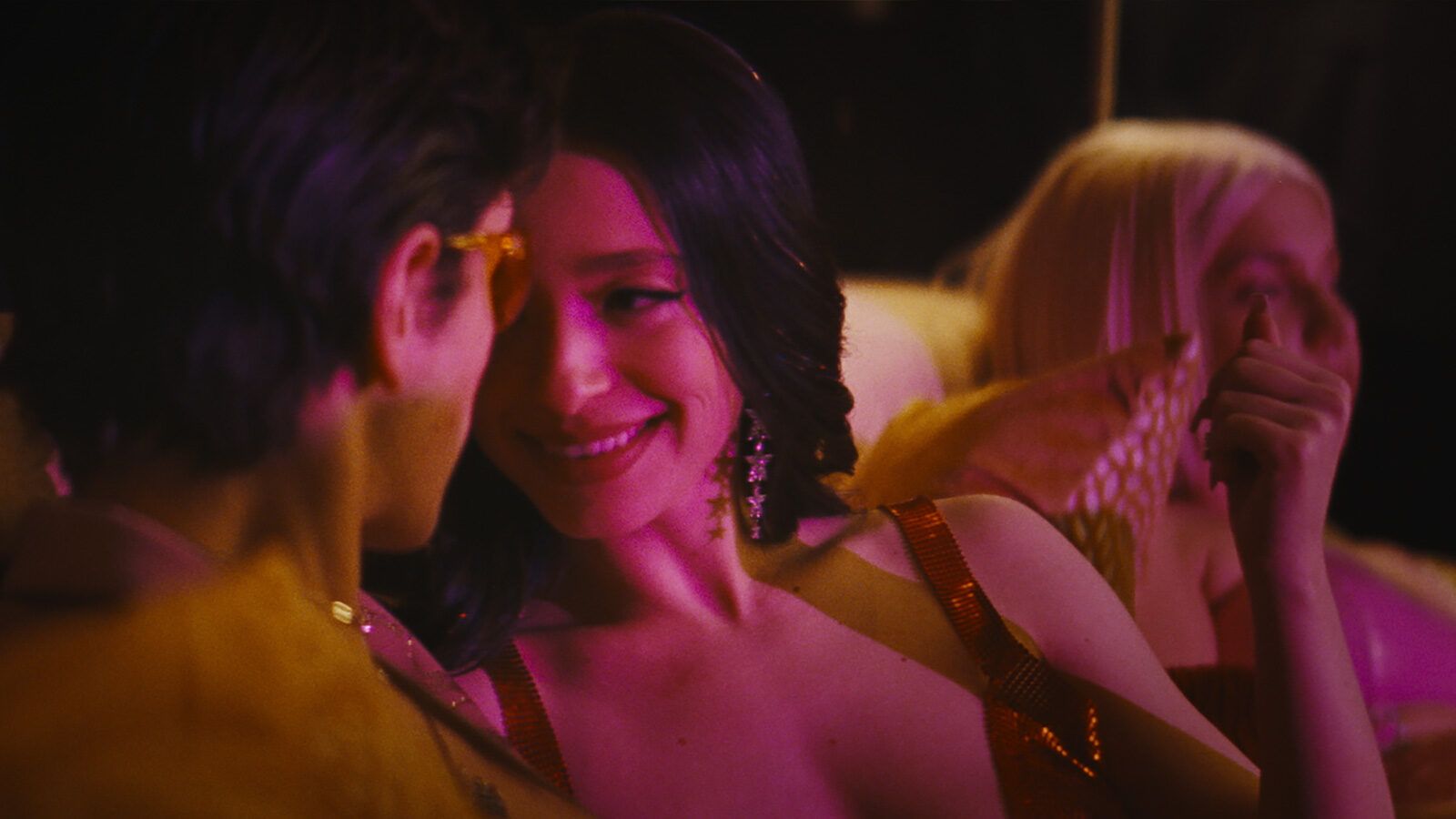 A woman with dark hair smiles at a man, illuminated by pink and orange lights in a club, with another person blurred behind them.