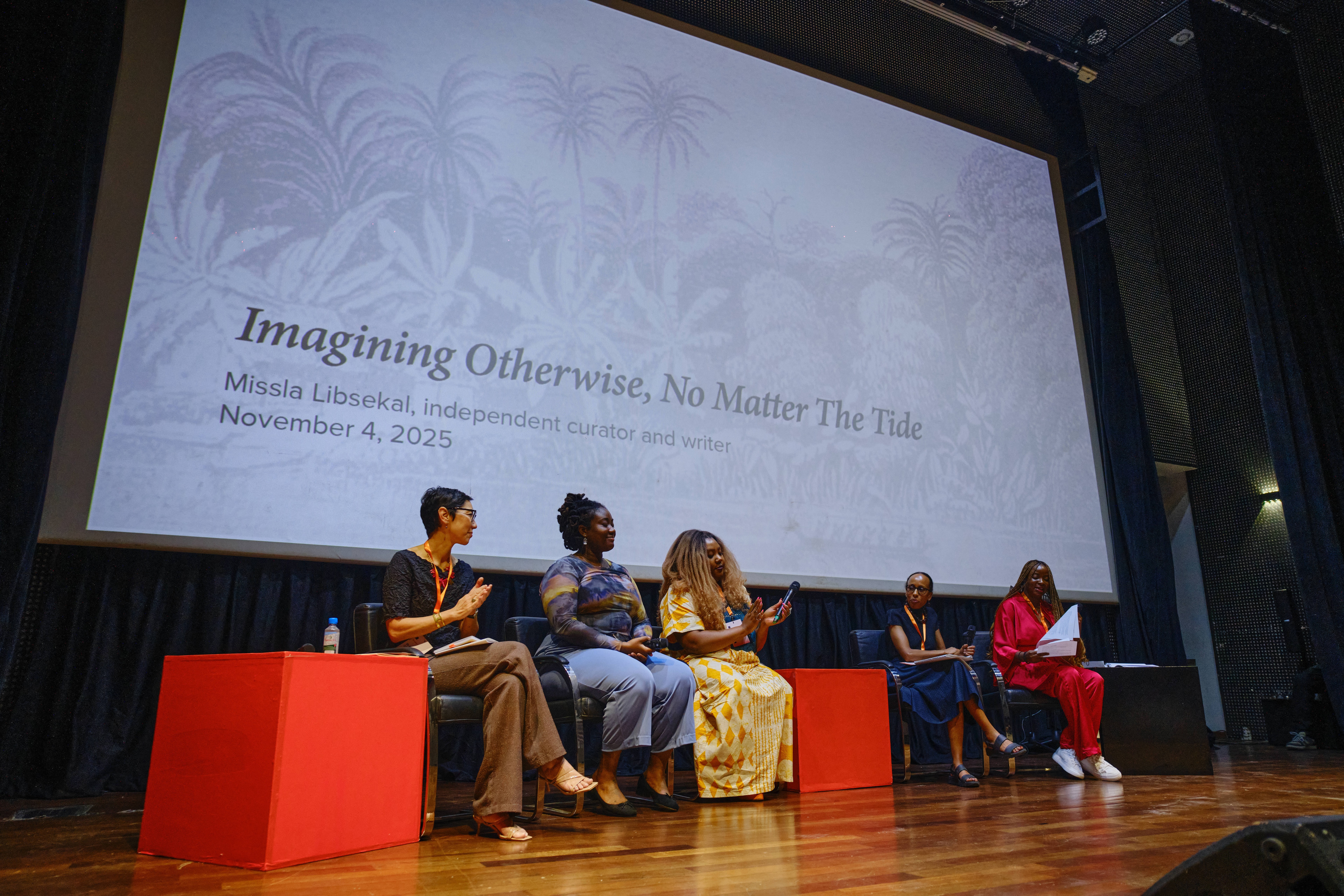 A panel of five women on a stage with "Imagining Otherwise, No Matter The Tide" displayed on a large screen behind them.