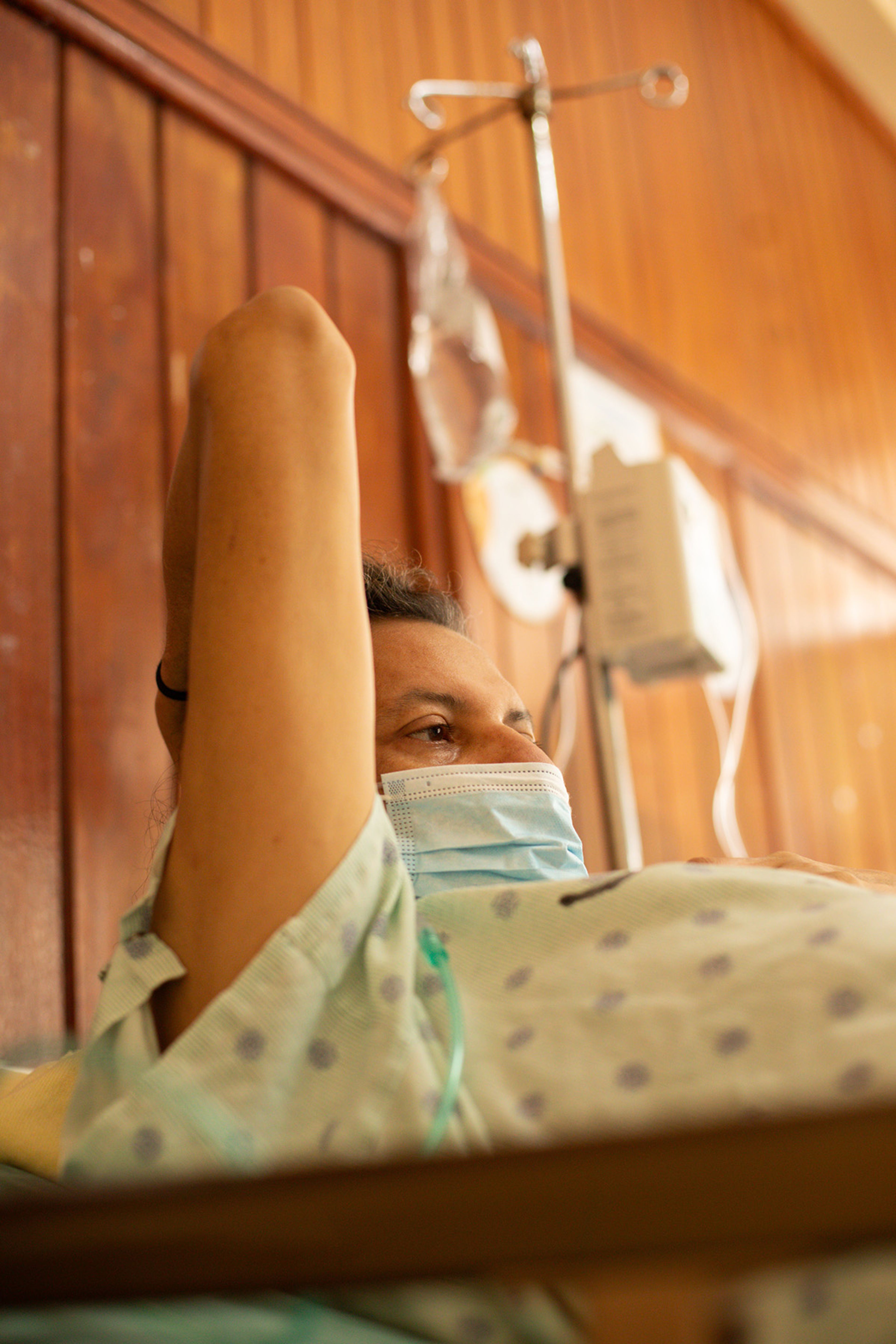A person in a hospital gown and mask lies in bed with an IV drip stand beside them.