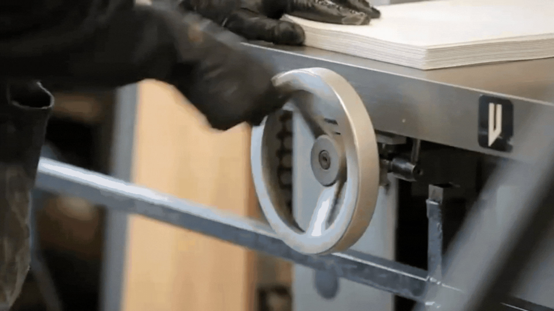 Gloved hands operating a machine with a large handwheel, next to a stack of papers.