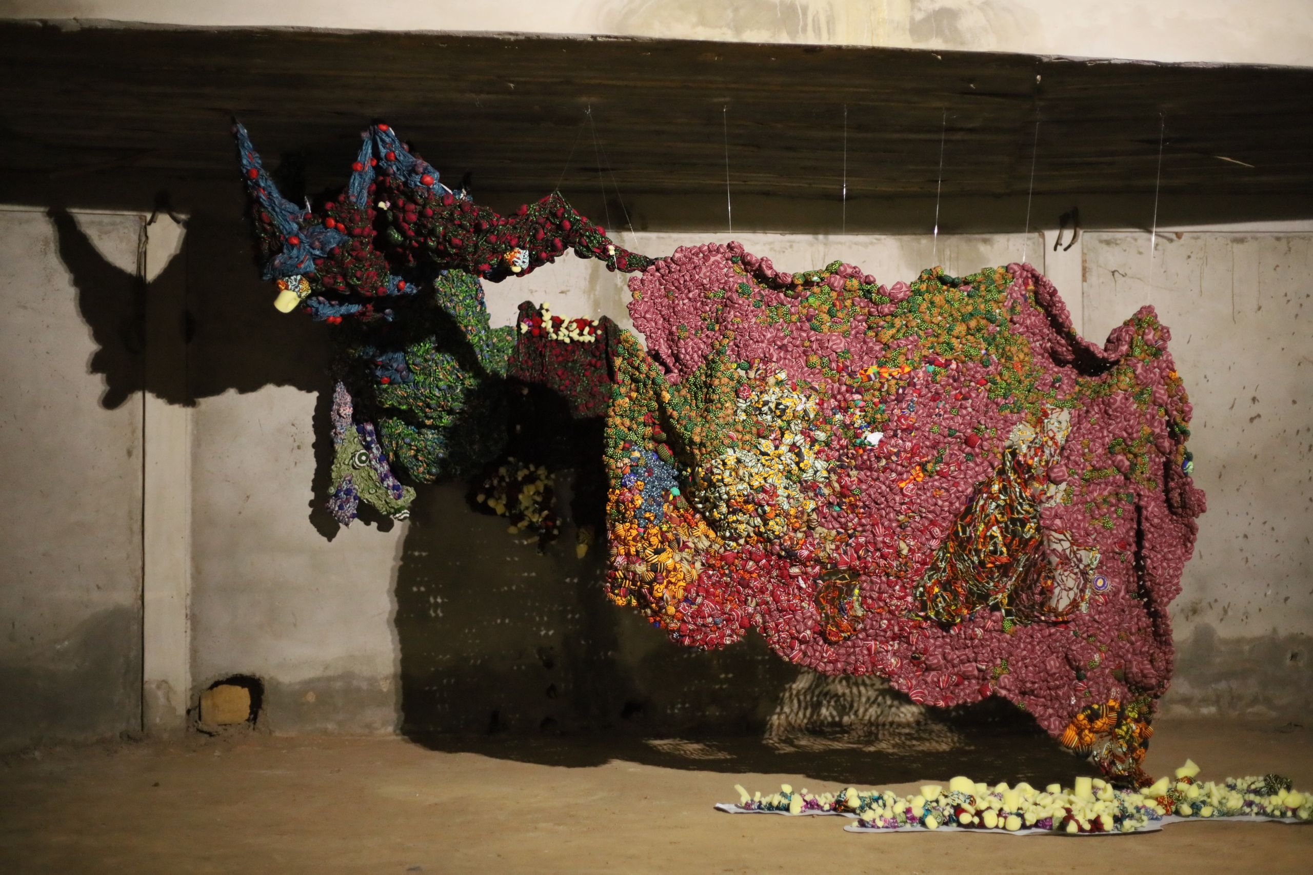An abstract sculpture made of thousands of colorful, bulbous fabric pieces, primarily pink, green, blue, and yellow, hangs in a raw concrete room.