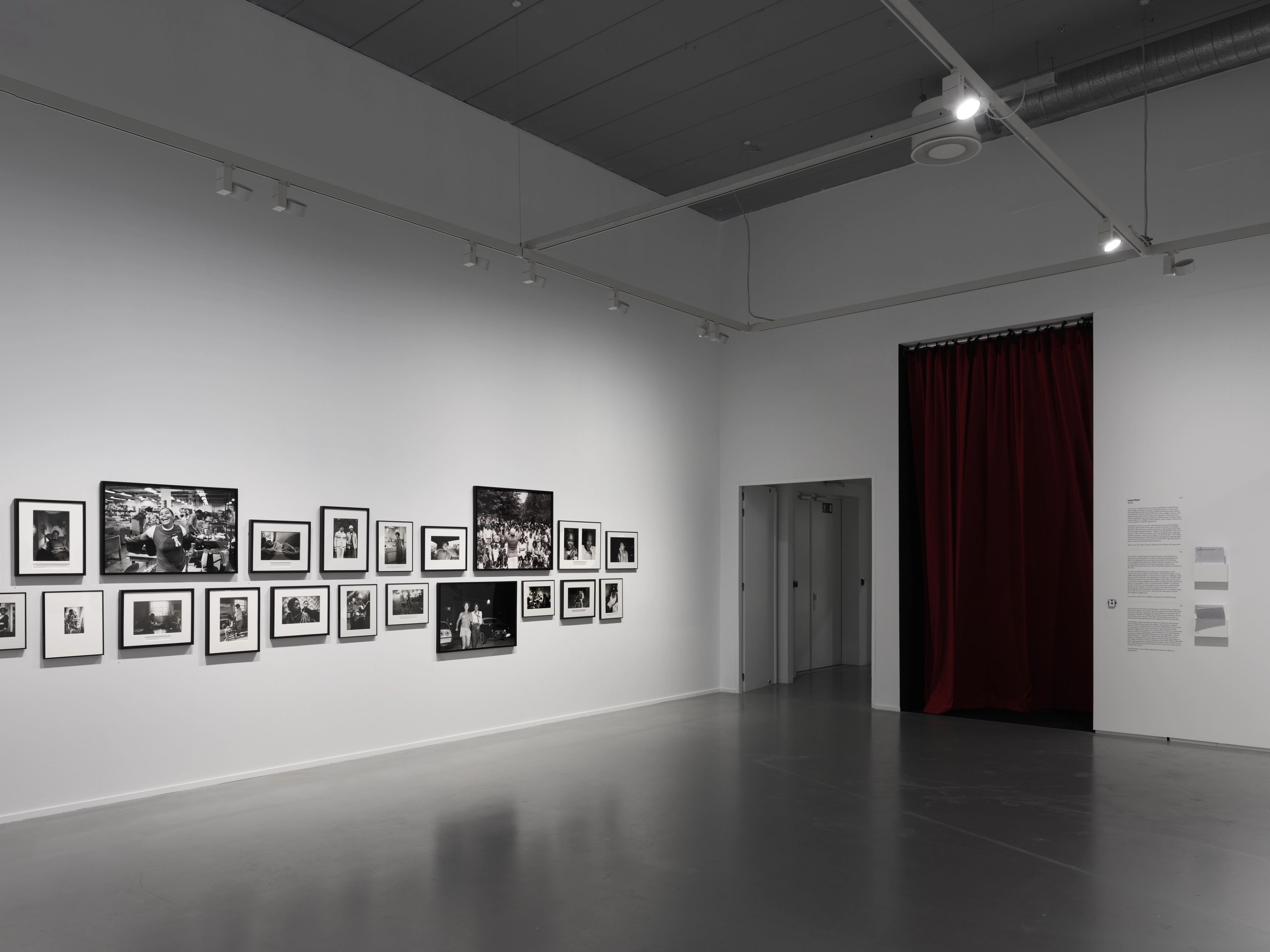 An art gallery displays black and white photographs on a white wall, with a dark gray floor and a red-curtained doorway.