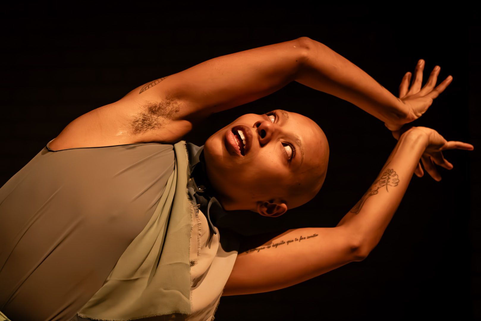 A person with a shaved head and visible armpit hair, wearing a sheer top, poses expressively with raised arms, looking up with an open mouth against a dark background.