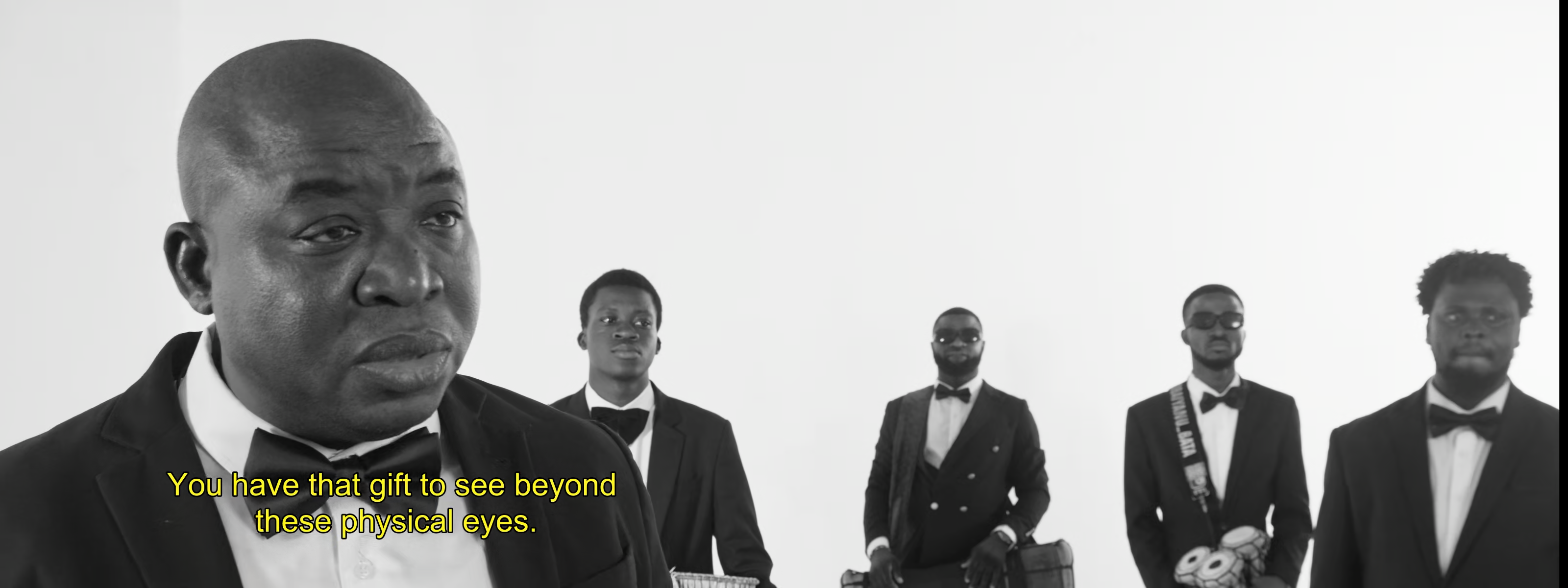 Black and white image of a man in a tuxedo speaking, with four men in tuxedos behind him. Subtitles read: "You have that gift to see beyond these physical eyes."