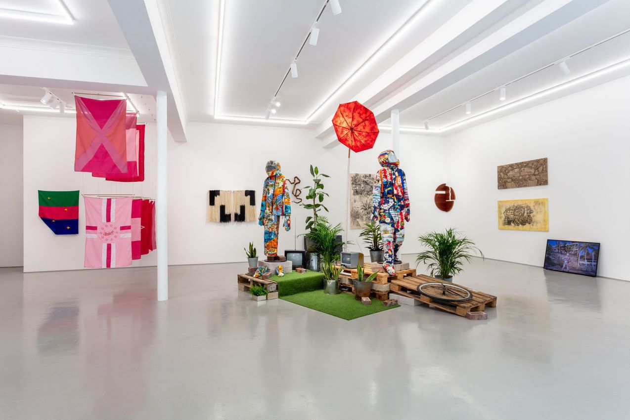 An art gallery displays a central installation with two colorful, collage-covered humanoid figures on artificial turf, surrounded by plants and old TVs. Flags hang on the left wall, and various artworks are on the right.