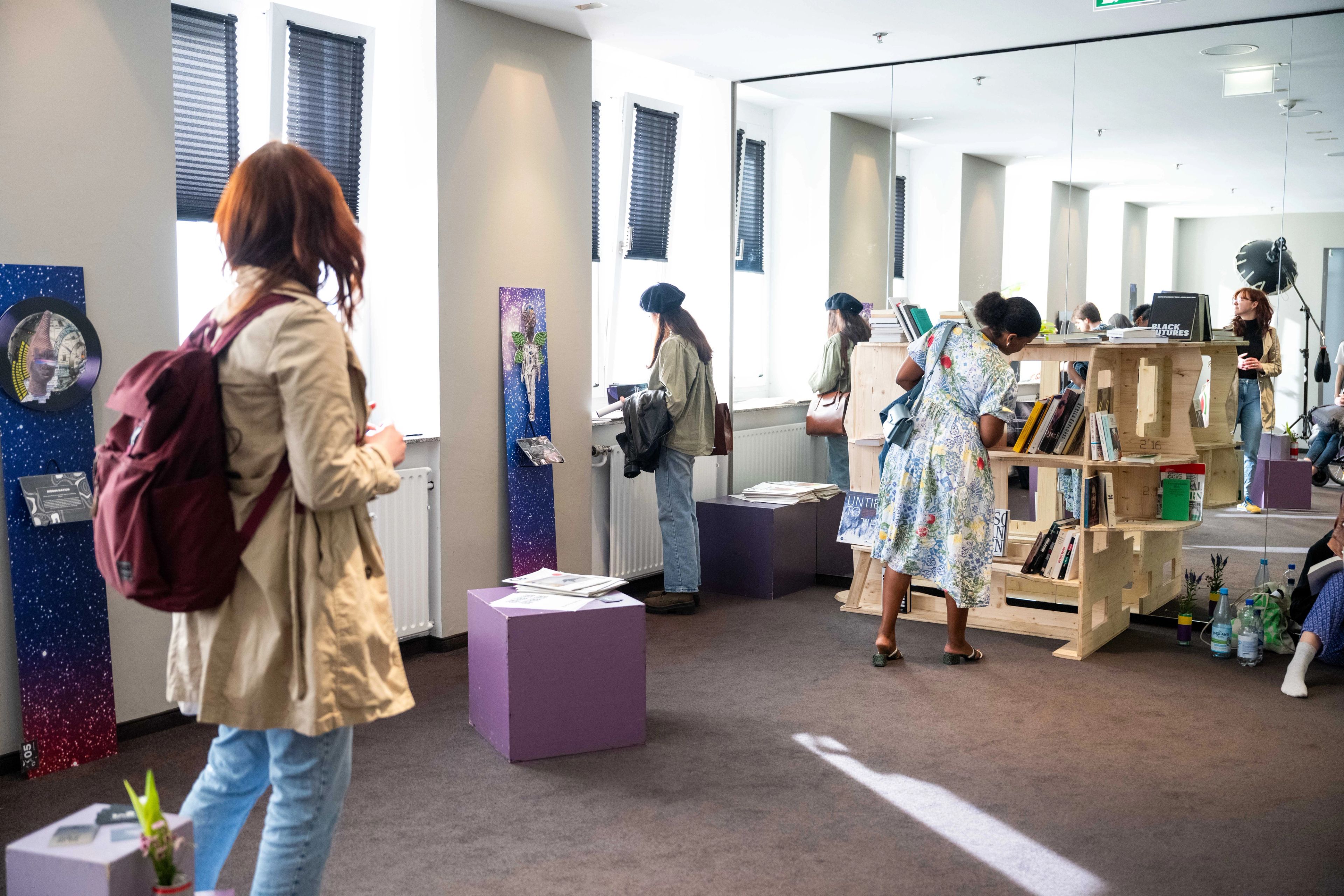 Visitors explore an indoor exhibition with galaxy-themed art displays, a wooden bookshelf, and other exhibits, reflected in a large mirror wall.