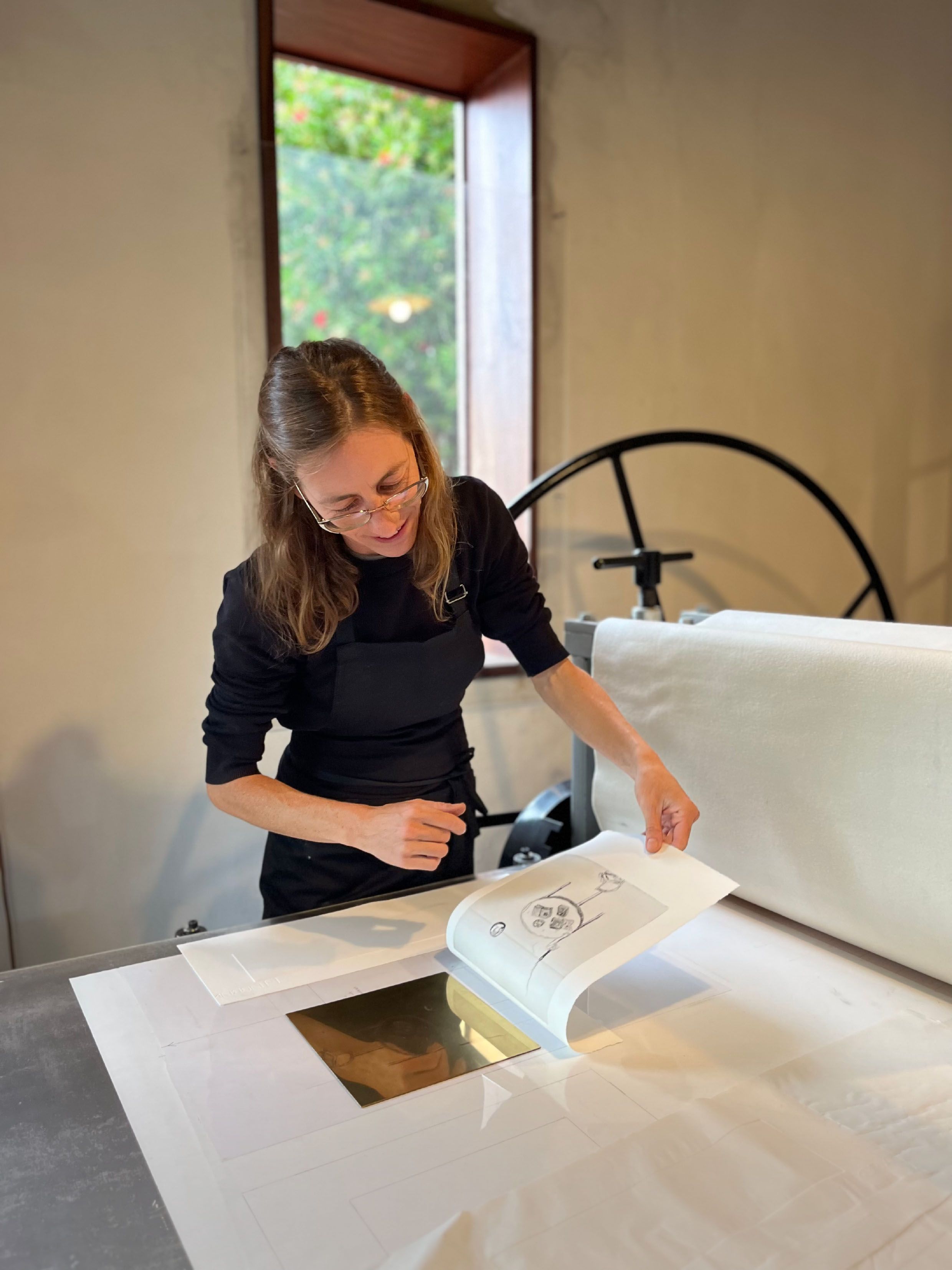 A smiling woman lifts a newly made print from a press in her art studio.