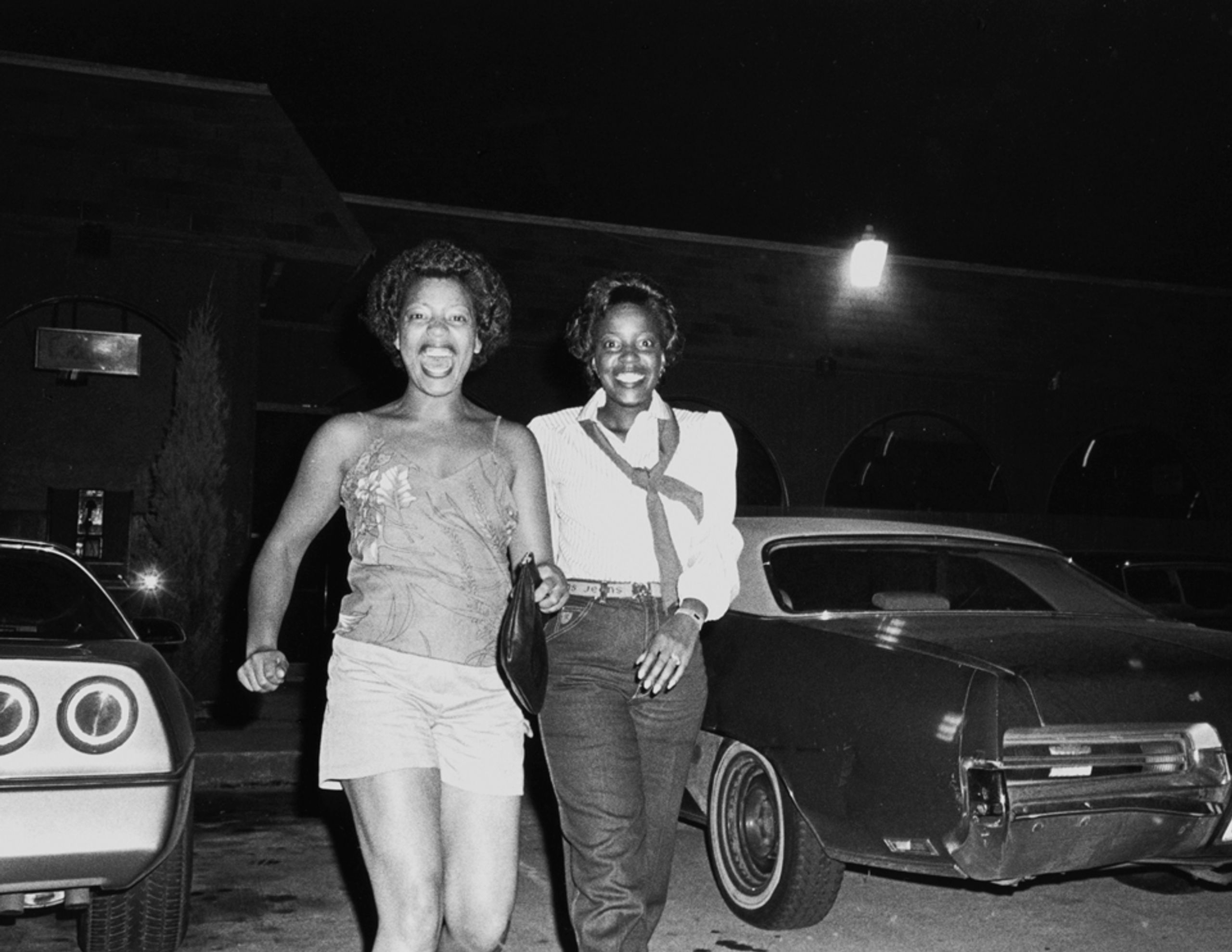 Black and white photo of two Black women smiling at night between cars, one sticking her tongue out.