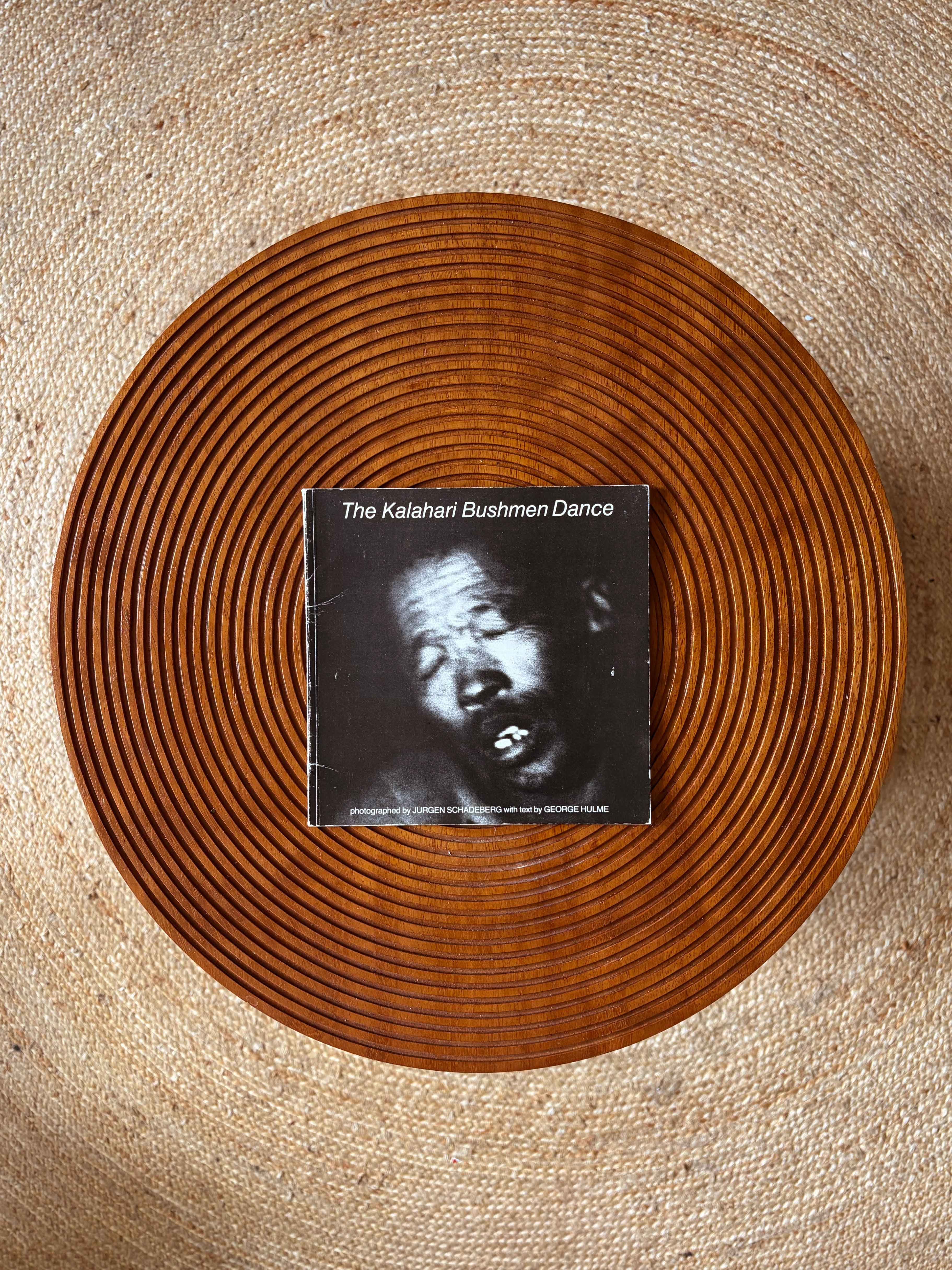 A book titled "The Kalahari Bushmen Dance" with a black and white photo of a man on its cover, rests on a round, ridged wooden tray, on a woven jute rug.