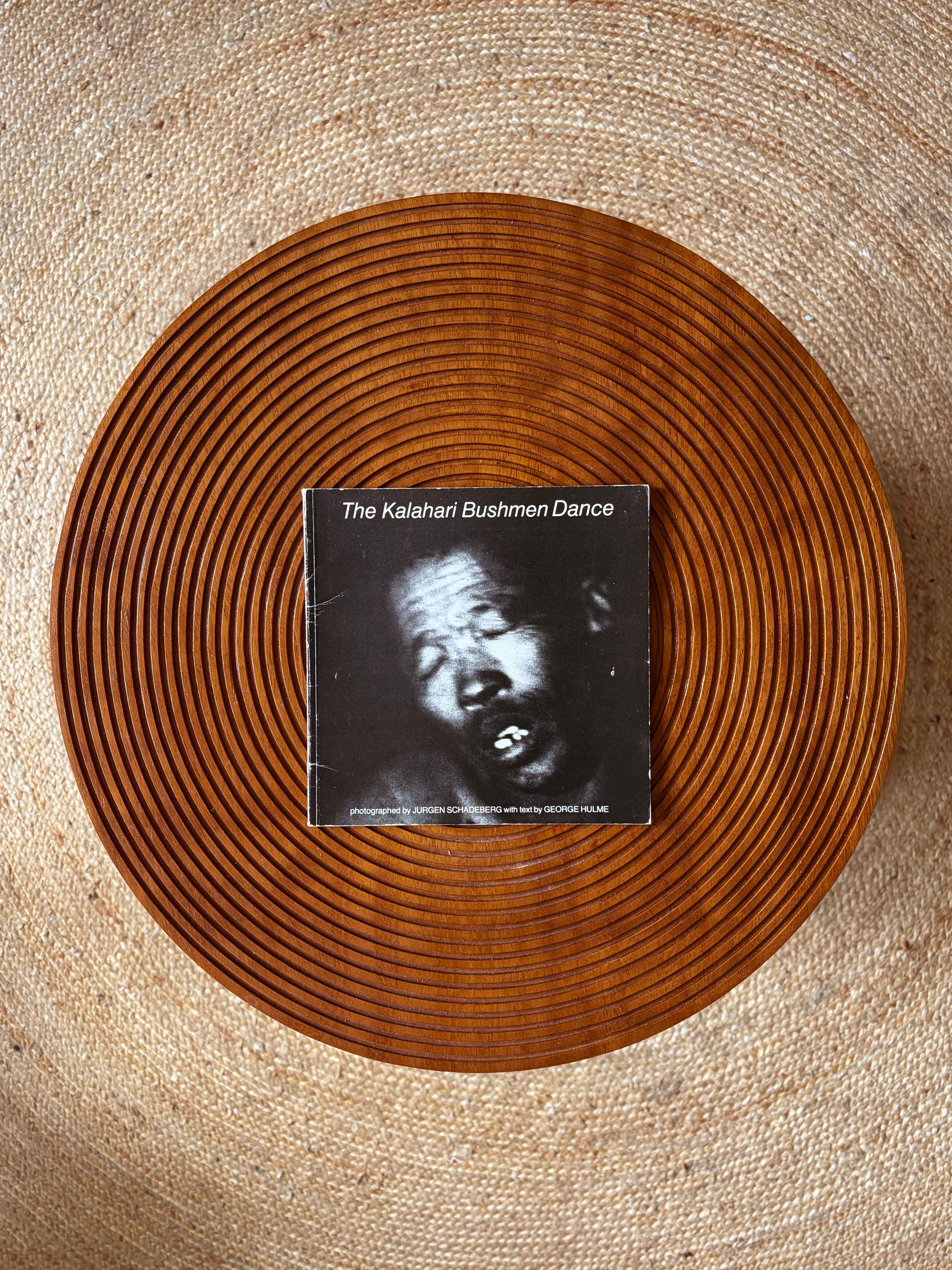A book titled "The Kalahari Bushmen Dance" with a black and white photo of a man on its cover, rests on a round, ridged wooden tray, on a woven jute rug.