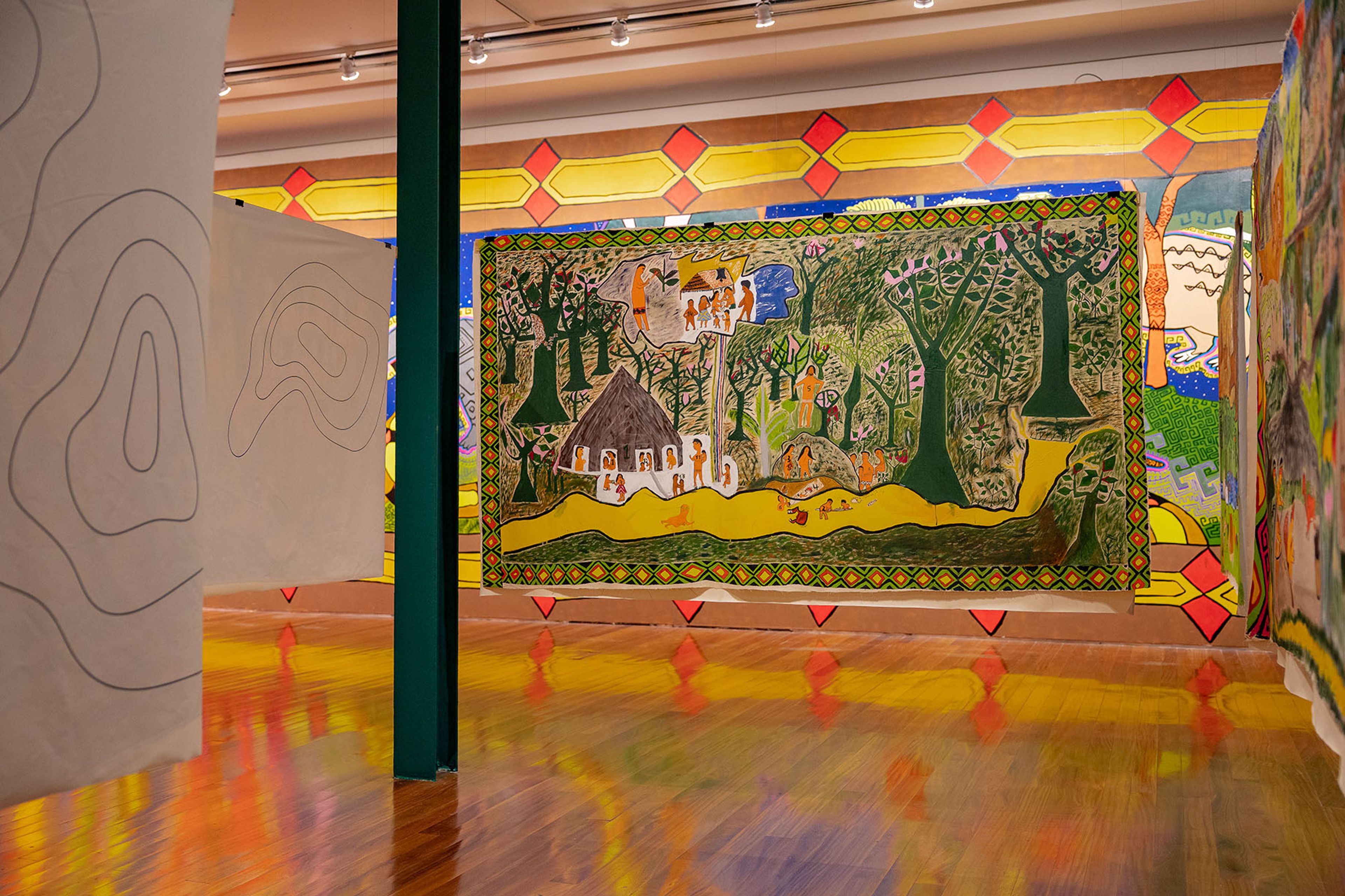 Colorful art exhibition with a large folk art tapestry depicting a village, trees, and people, flanked by patterned artworks and a reflective wooden floor.
