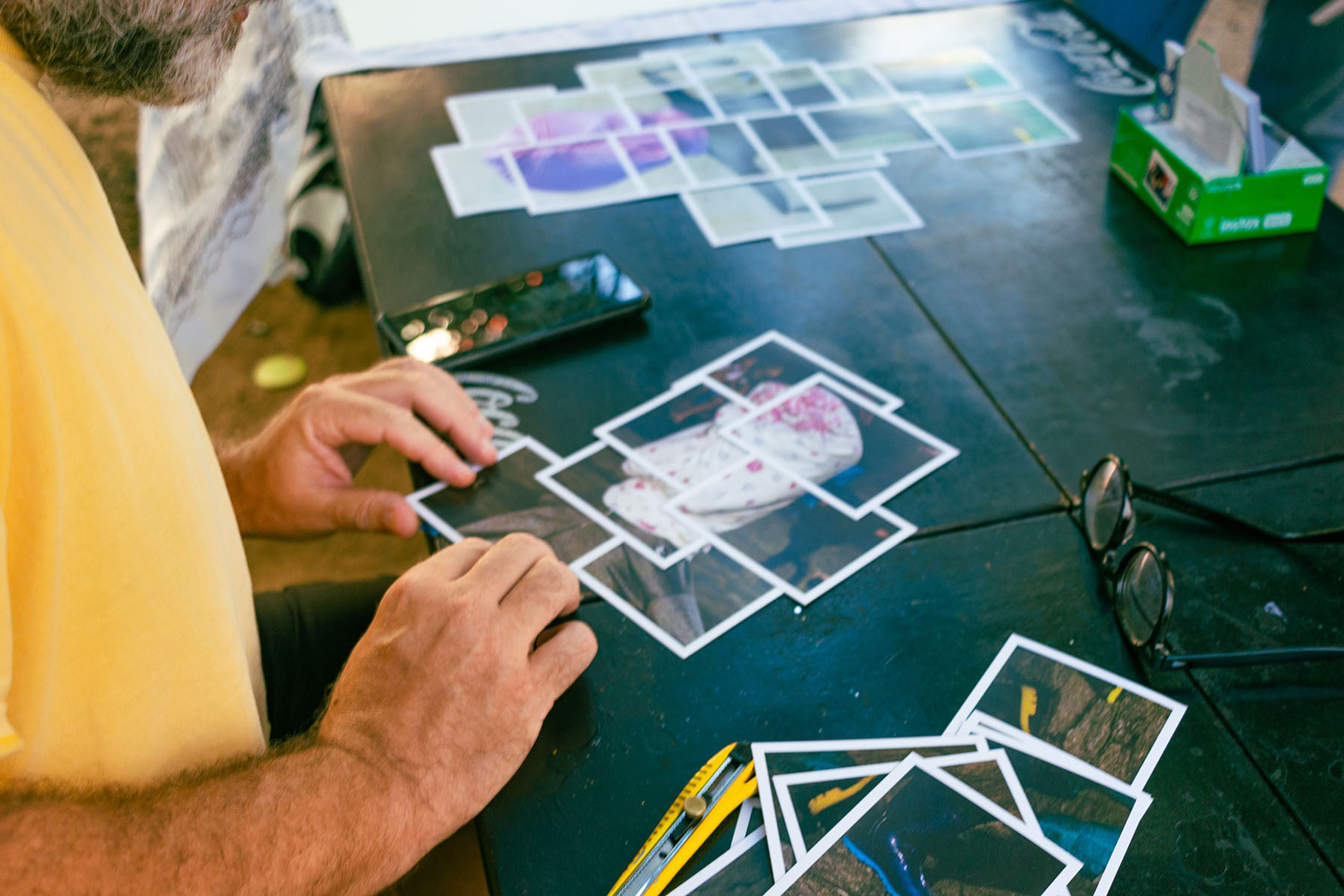 A person's hands arrange small printed photos on a dark table, with more photos, a phone, and glasses nearby.