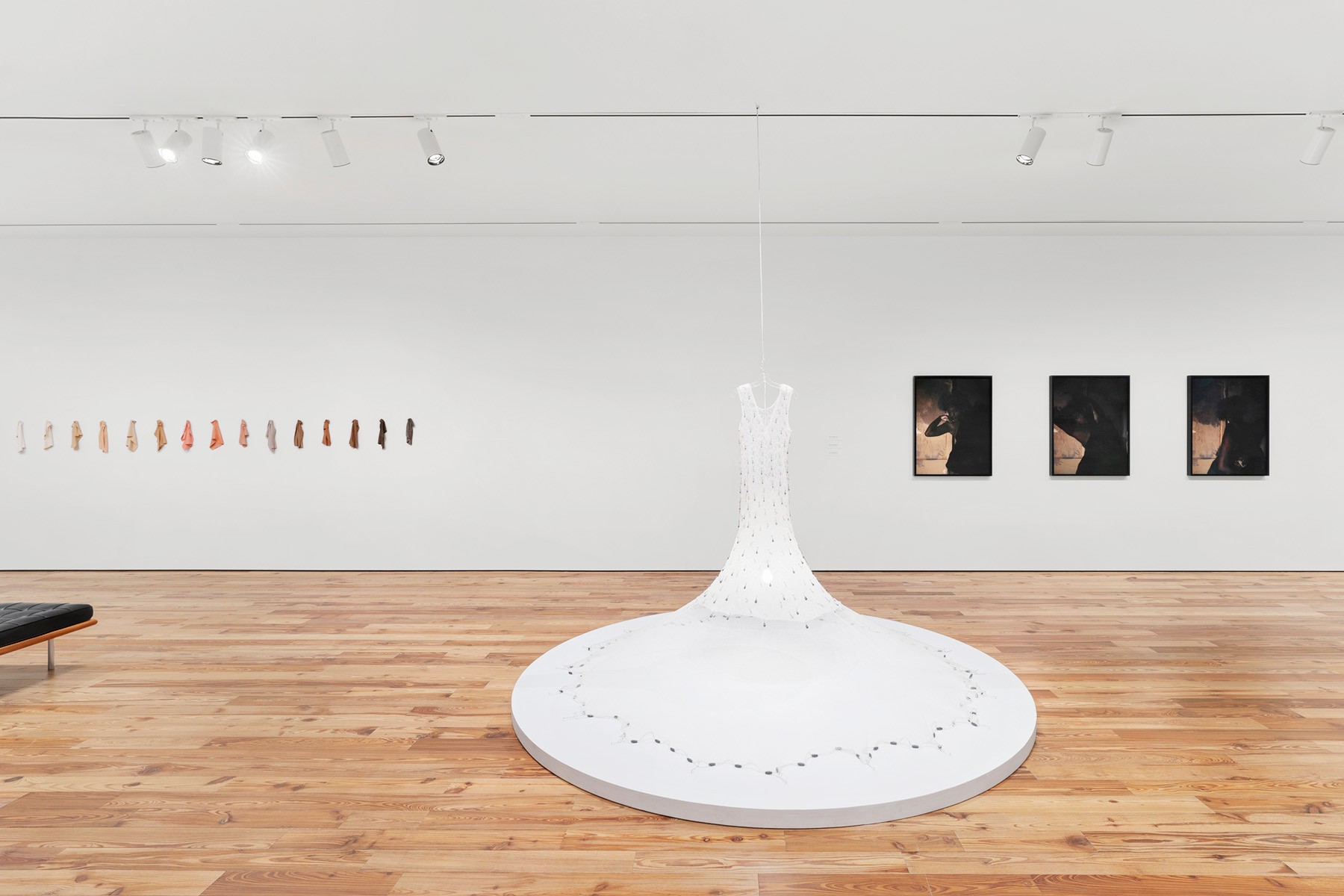 Art gallery with a central illuminated white dress sculpture, a row of small flesh-toned objects, and three dark-toned paintings.