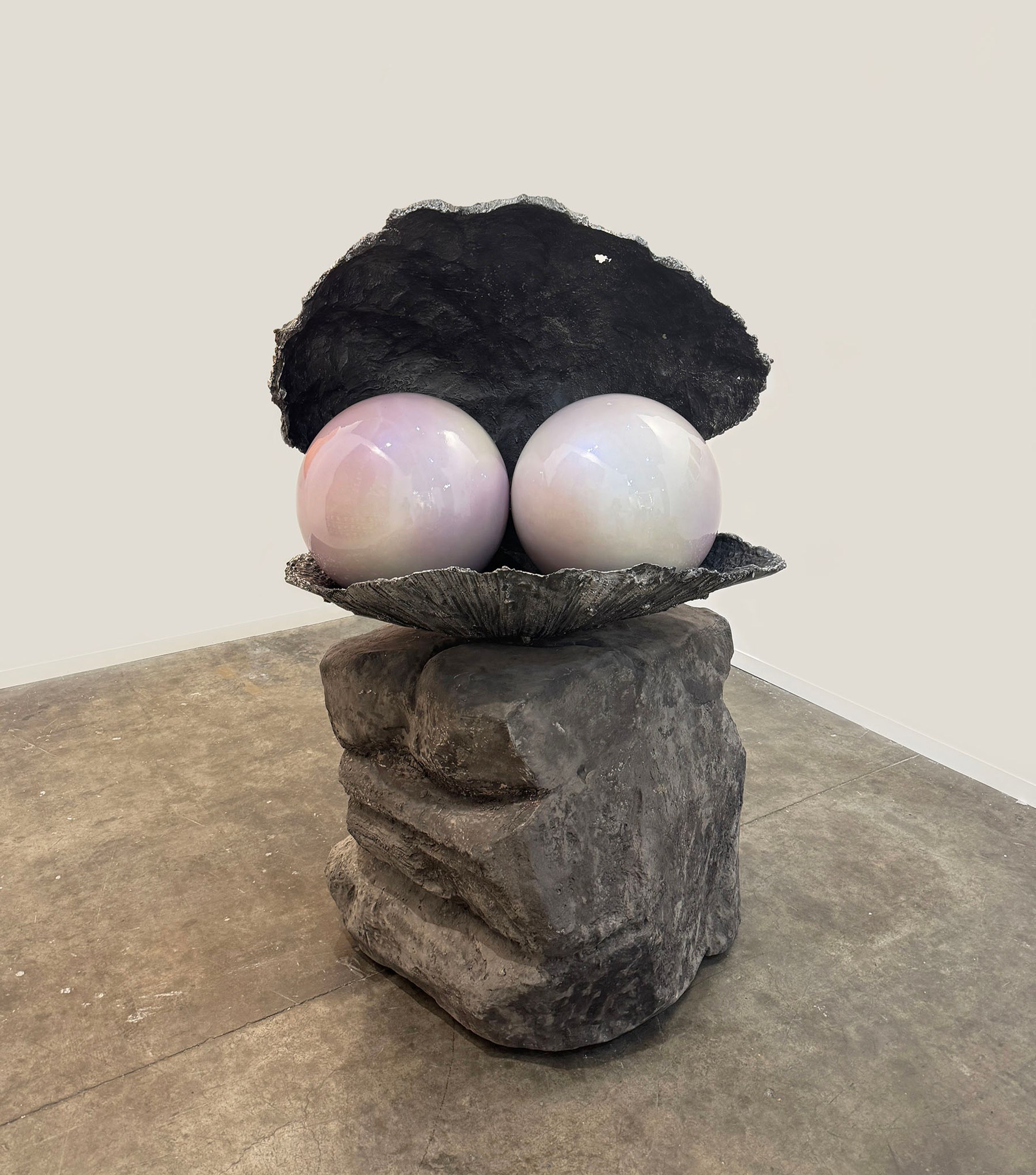 A sculpture of two large, iridescent pearls inside an open, dark oyster shell, resting on a stacked stone base.