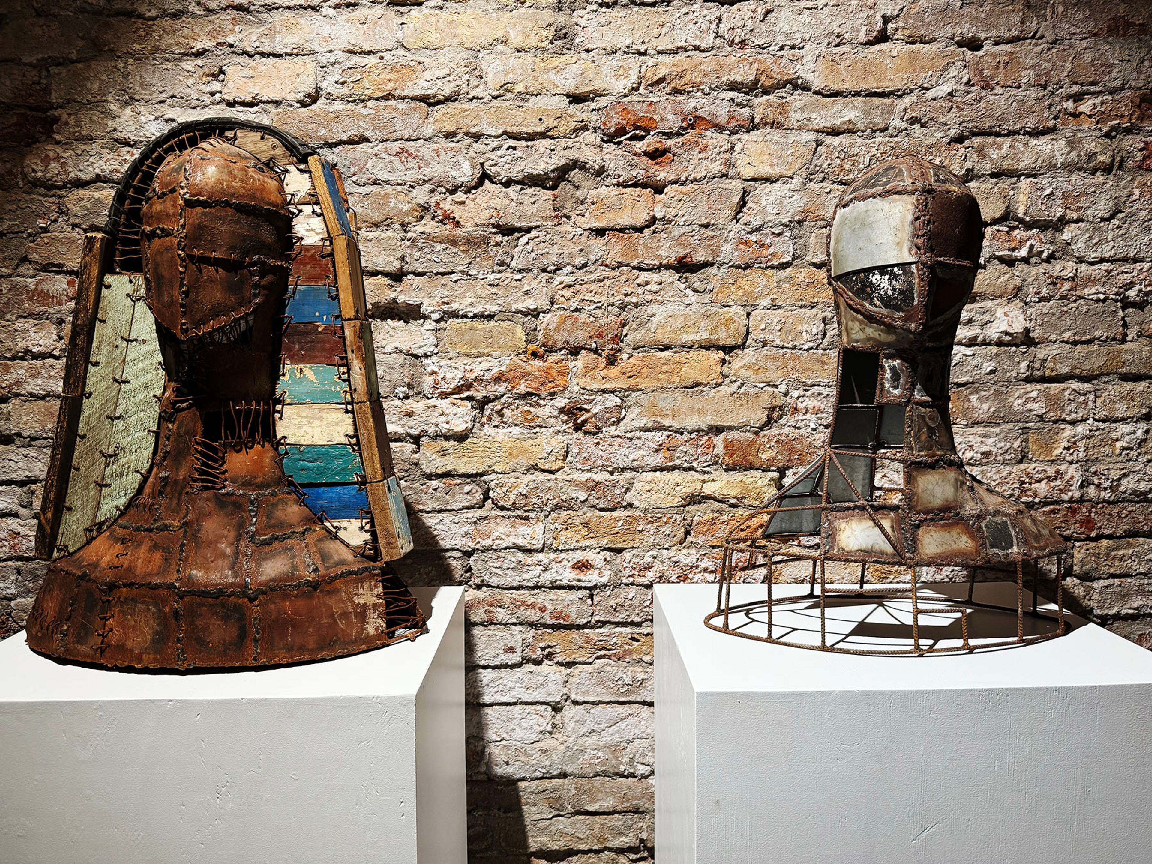 Two artistic busts made of rusted metal and salvaged materials. One features colorful side panels, the other an open frame. On white pedestals against a brick wall.