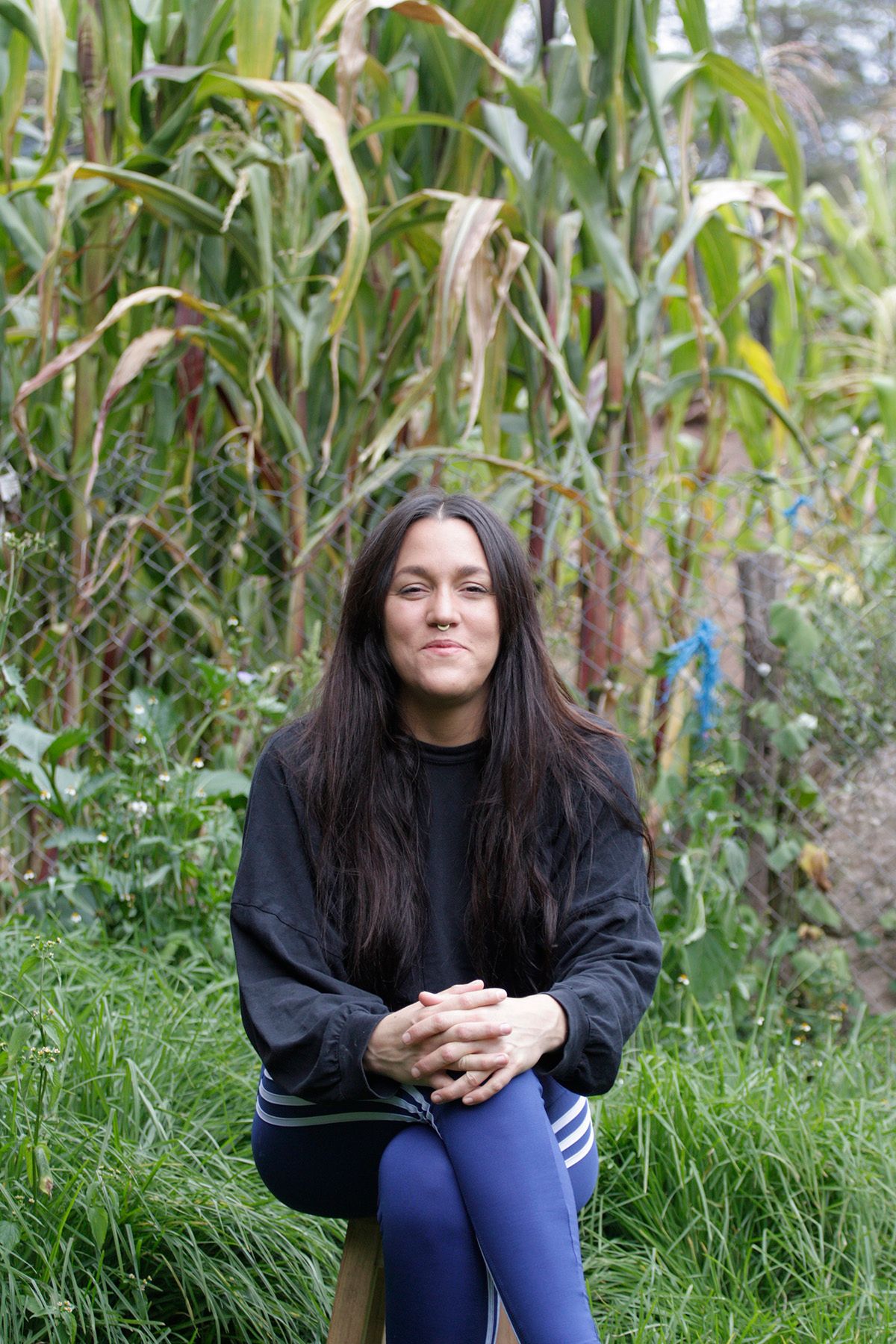 A woman with long dark hair and a nose ring sits on a stool in a garden with tall corn plants, looking at the camera with a faint smile.