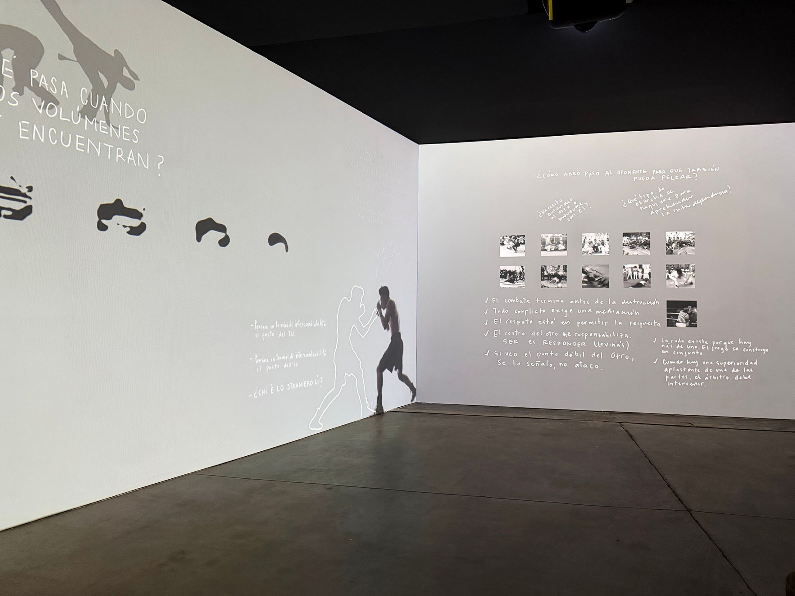 A dark room with white walls displaying projected art: a boxer's shadow, abstract shapes, a grid of photos, and handwritten Spanish text.