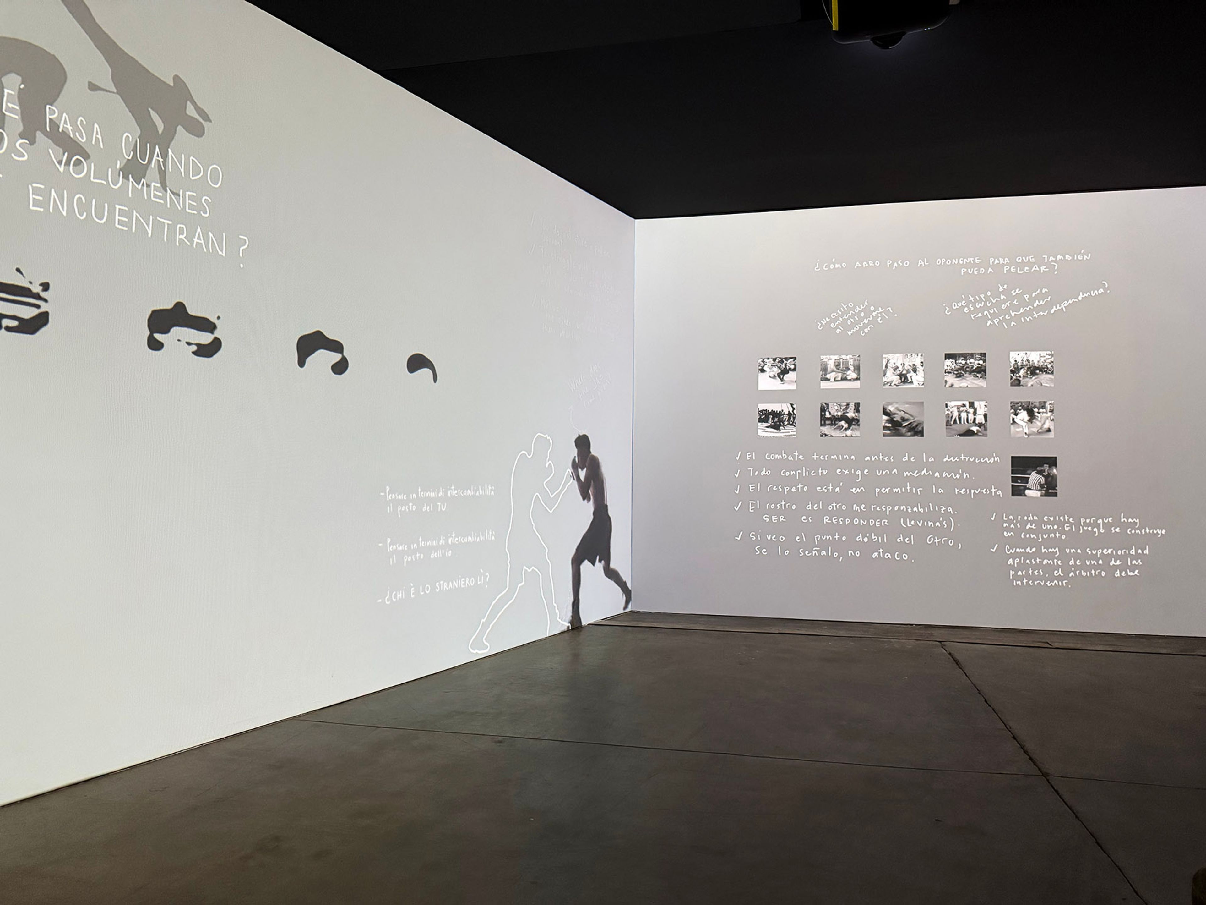 A dark room with white walls displaying projected art: a boxer's shadow, abstract shapes, a grid of photos, and handwritten Spanish text.