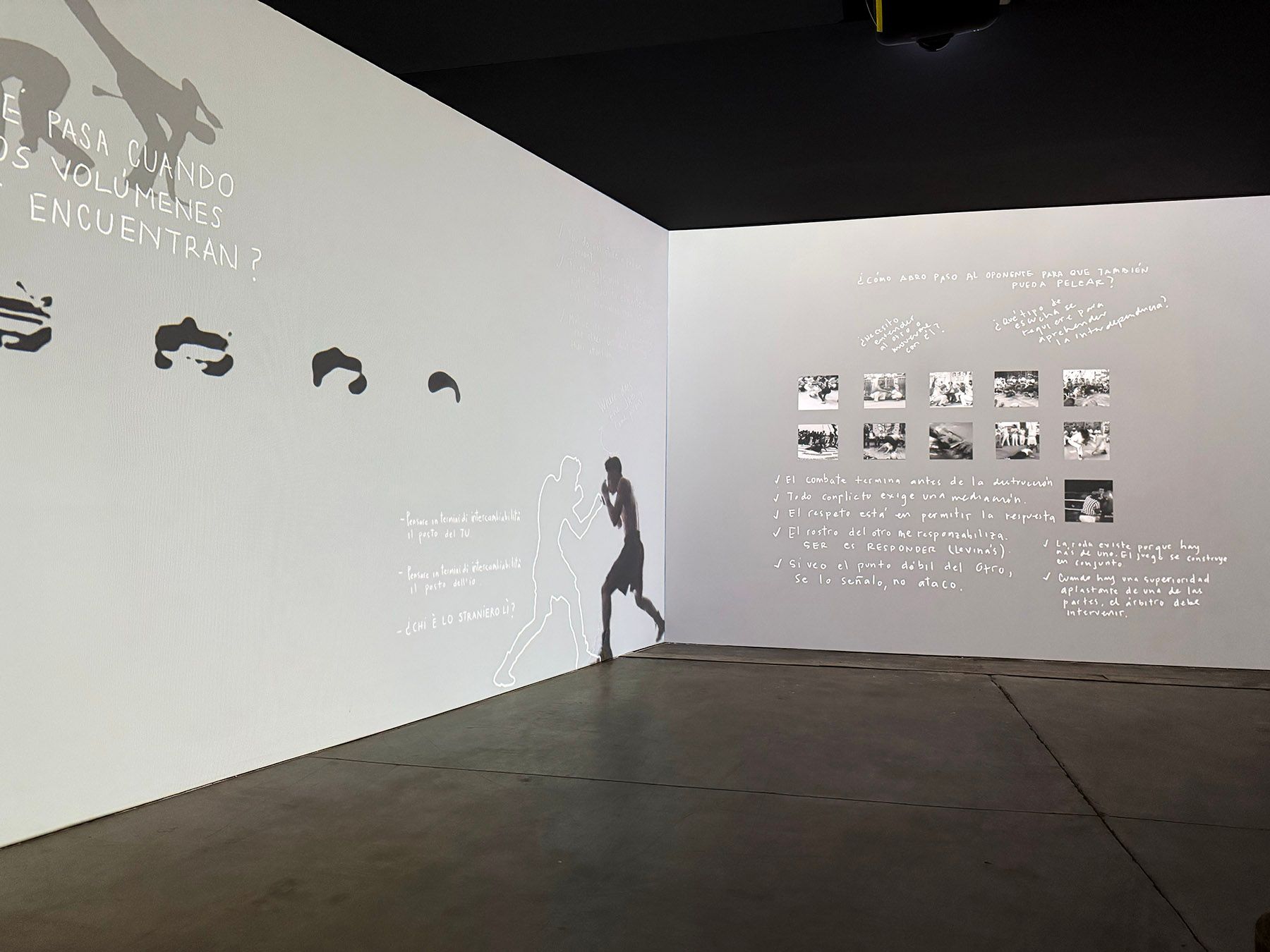 A dark room with white walls displaying projected art: a boxer's shadow, abstract shapes, a grid of photos, and handwritten Spanish text.
