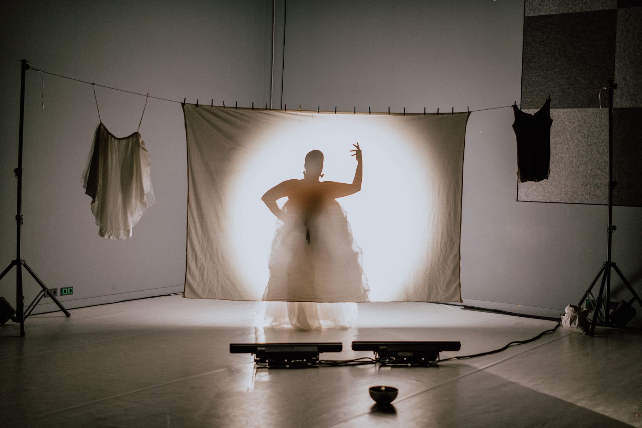 A person's silhouette, illuminated by a spotlight from behind, stands behind a white screen on a stage, flanked by white and black hanging garments.