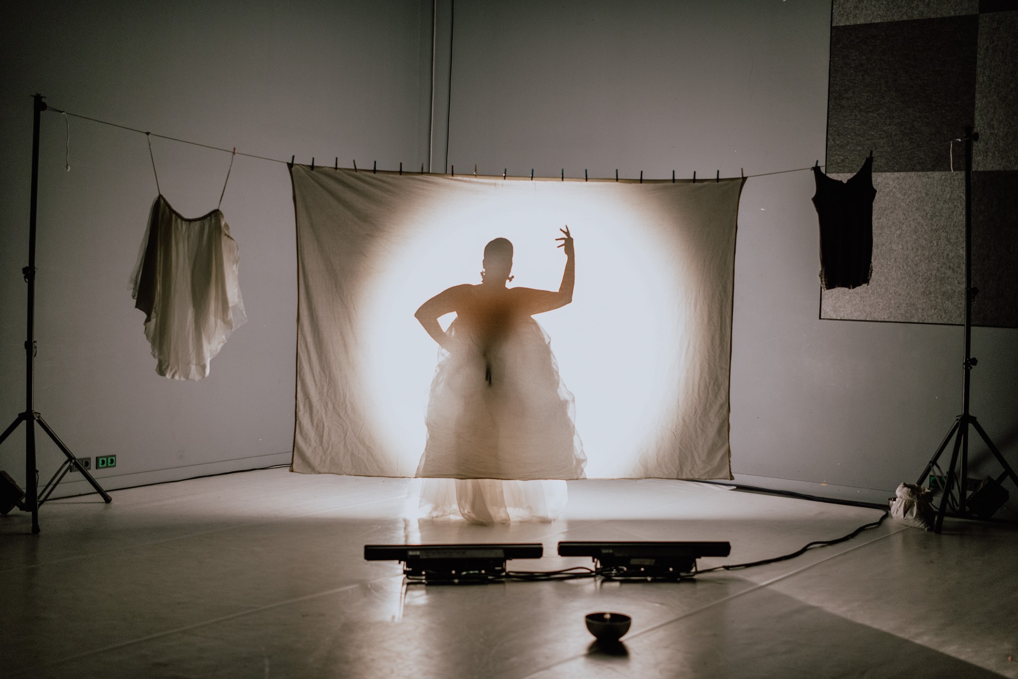 A person's silhouette, illuminated by a spotlight from behind, stands behind a white screen on a stage, flanked by white and black hanging garments.