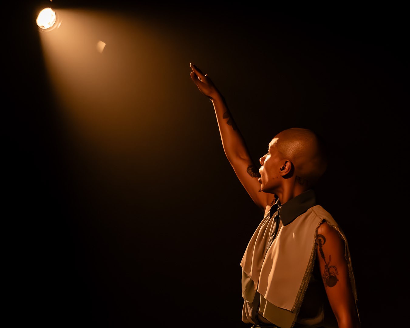 A bald person with dark skin raises an arm towards a bright spotlight, looking up with an open mouth against a dark background.