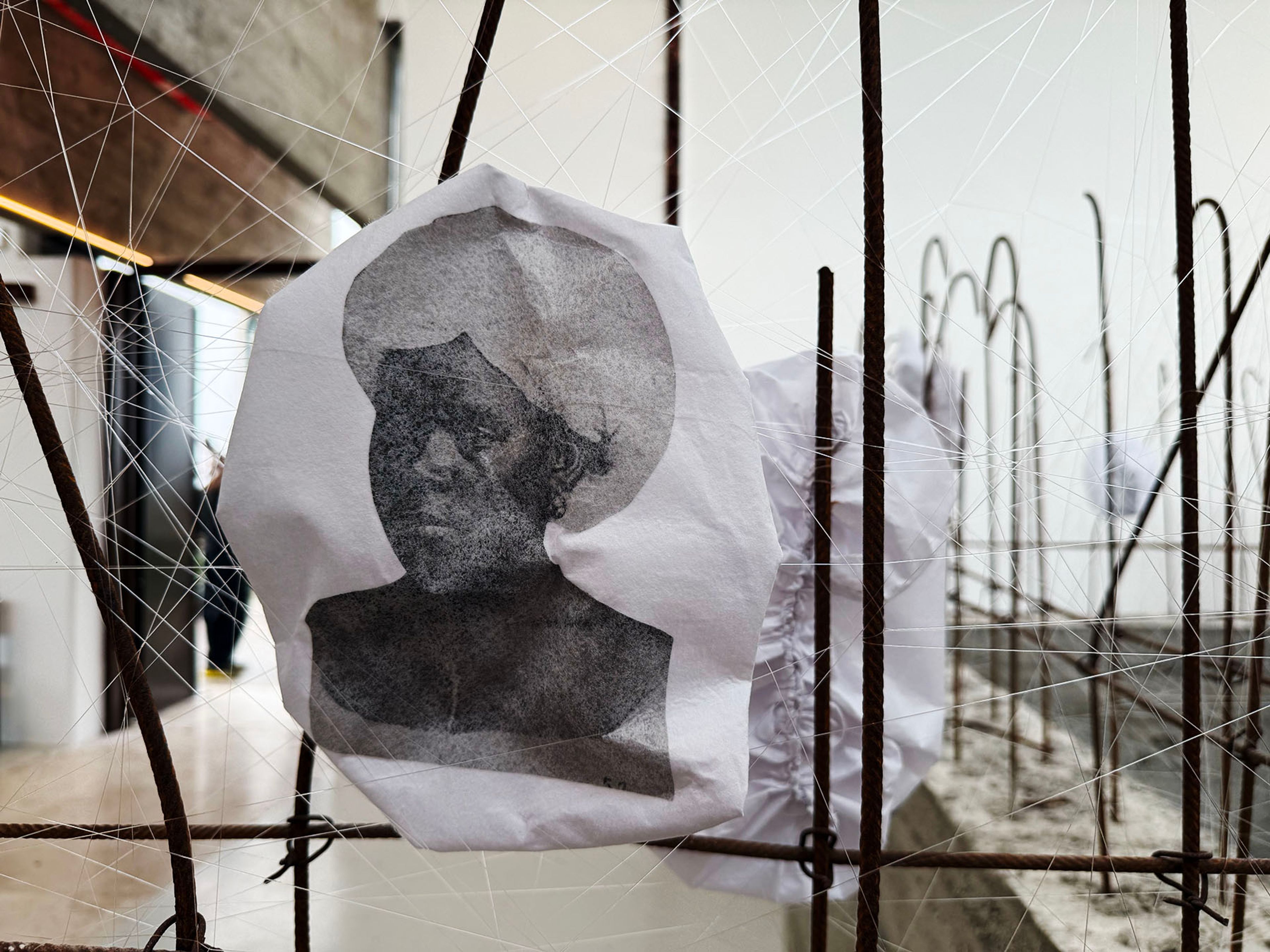 A black and white portrait on crumpled paper hangs among rusty rebar and white strings.