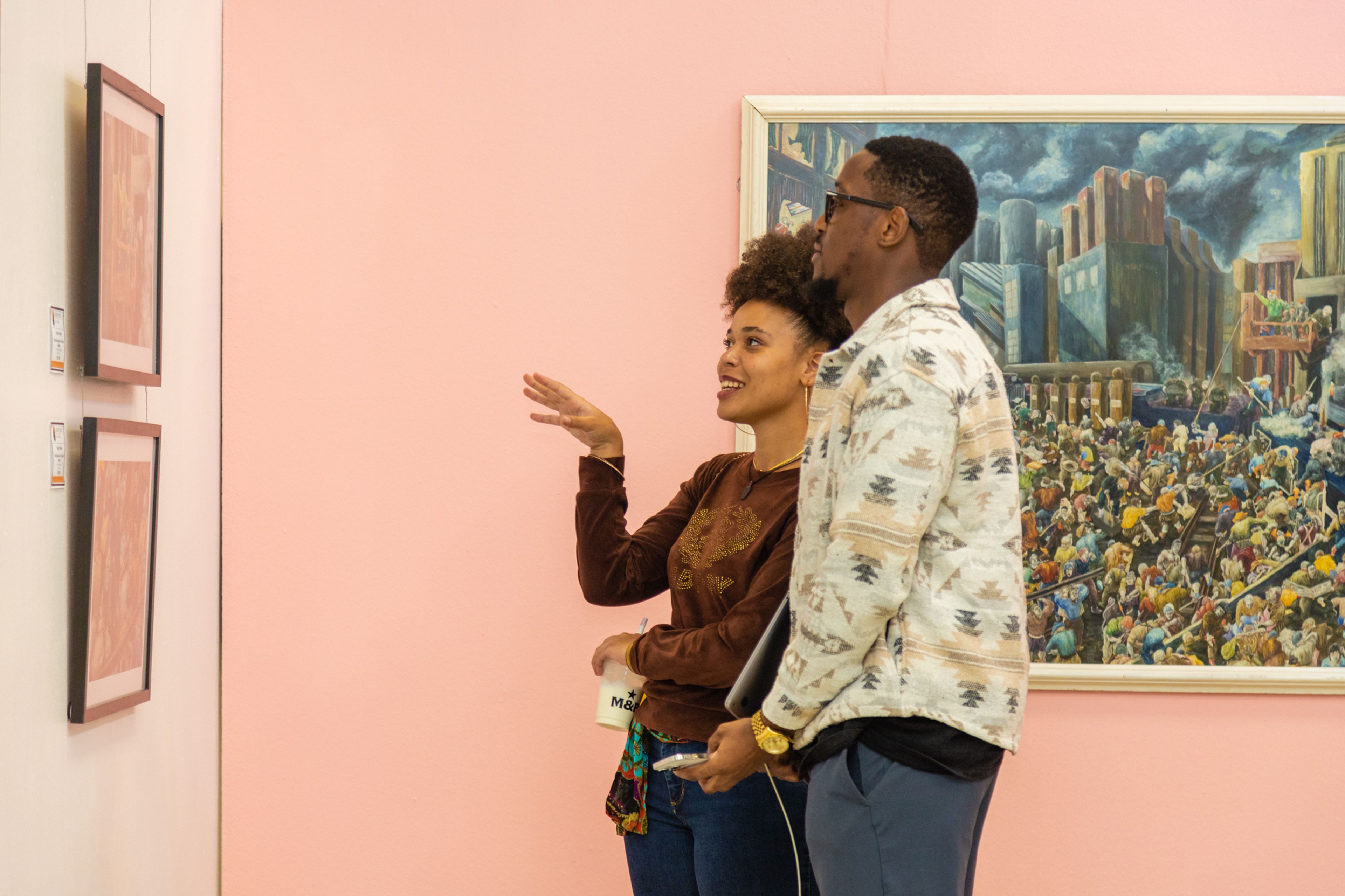 A woman gesturing and a man looking at a large painting of a crowd and city buildings in an art gallery.