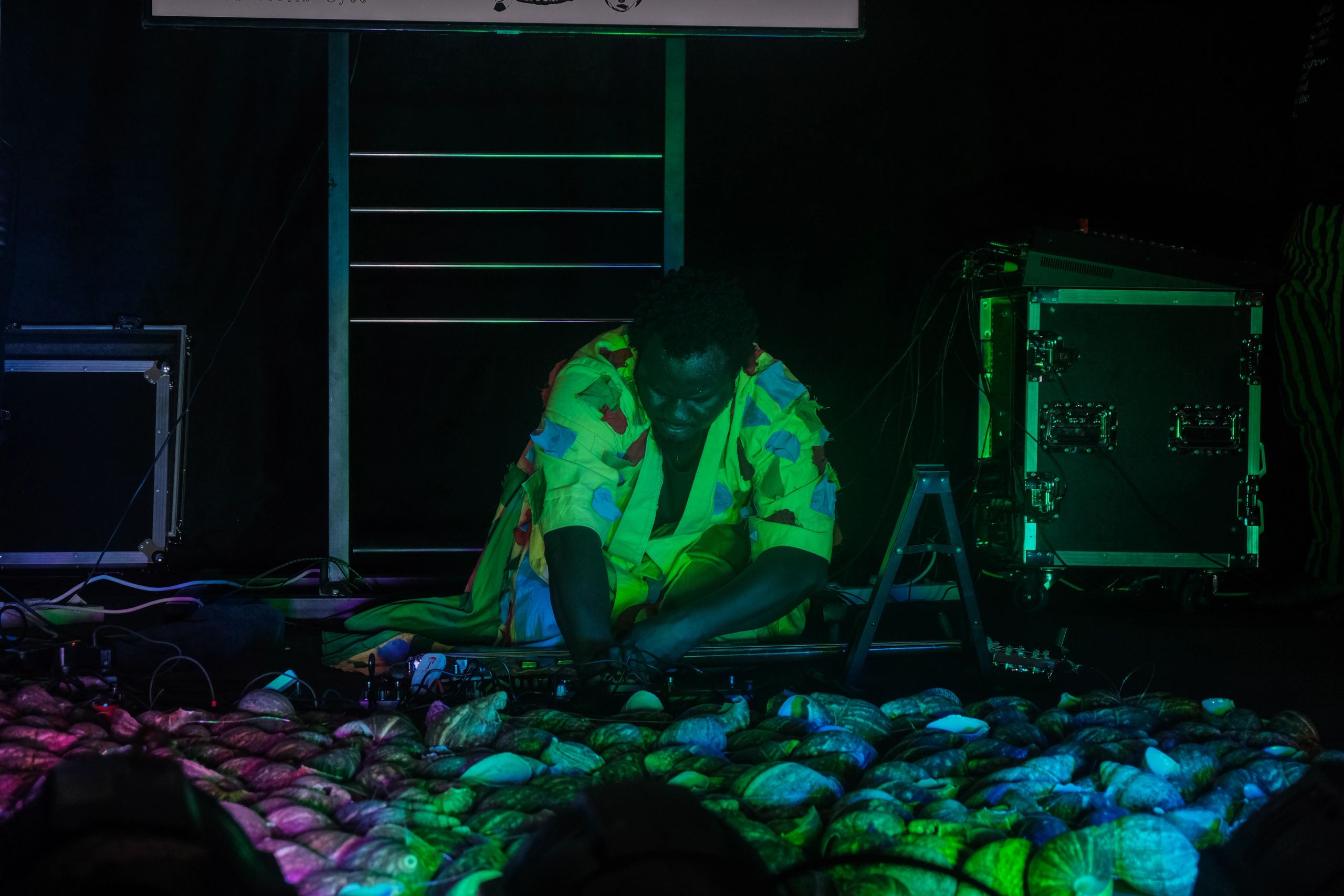 A person in a bright green patterned outfit kneels on a dark stage, interacting with a sprawling arrangement of shells illuminated by purple and green light.