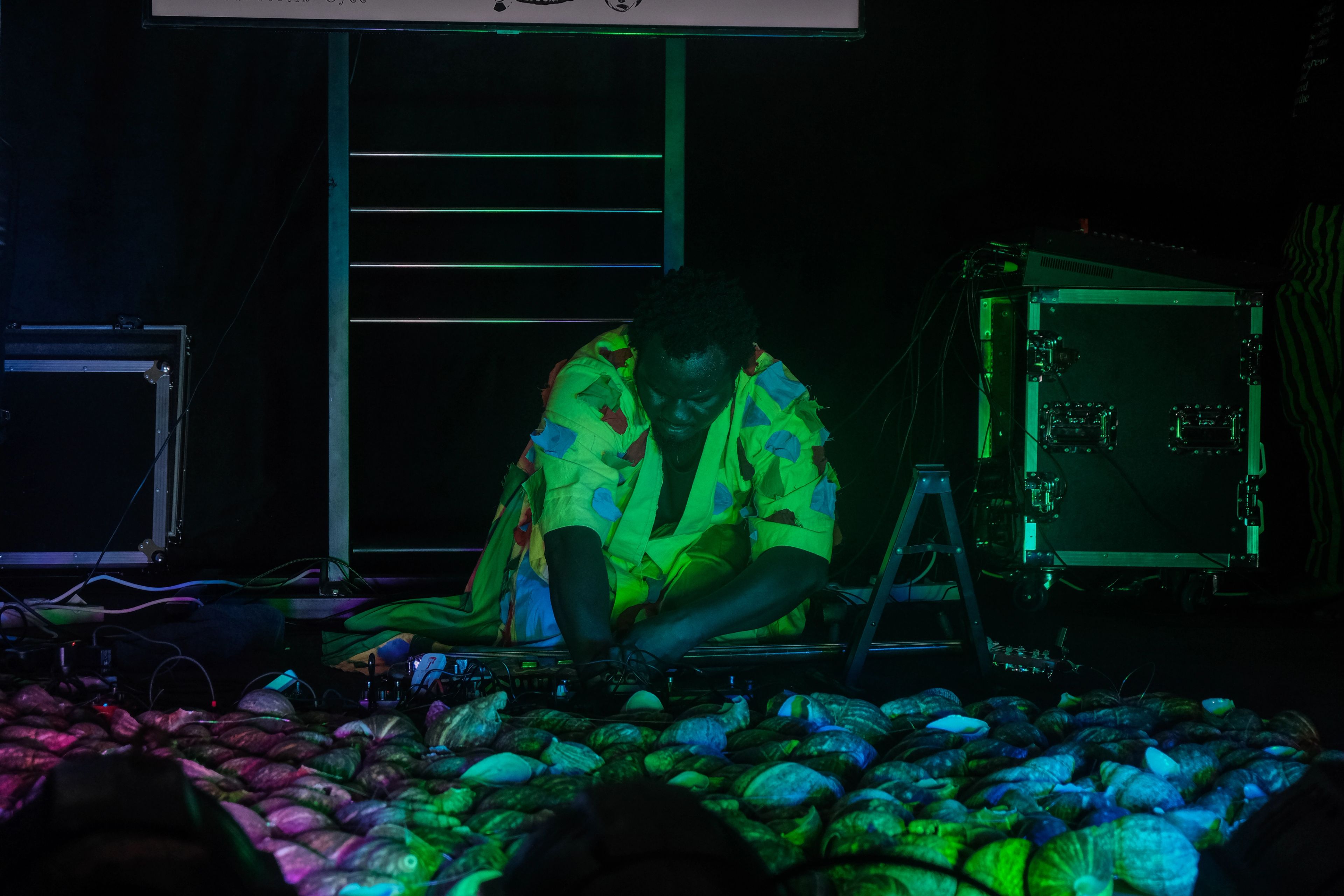A person in a bright green patterned outfit kneels on a dark stage, interacting with a sprawling arrangement of shells illuminated by purple and green light.